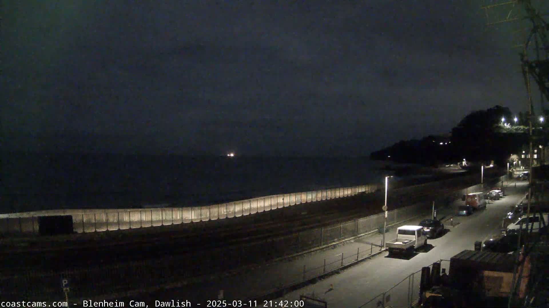 A nighttime coastal scene shows a dark sea, a long seawall with lights, a street with parked cars, and a hill in the background under a cloudy sky.