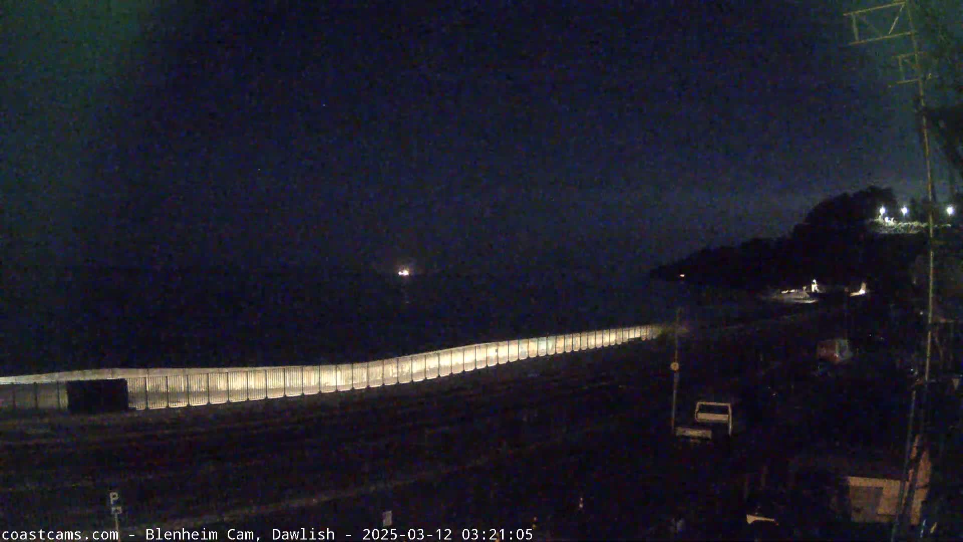 A long, illuminated fence runs along a dark coastline at night.
