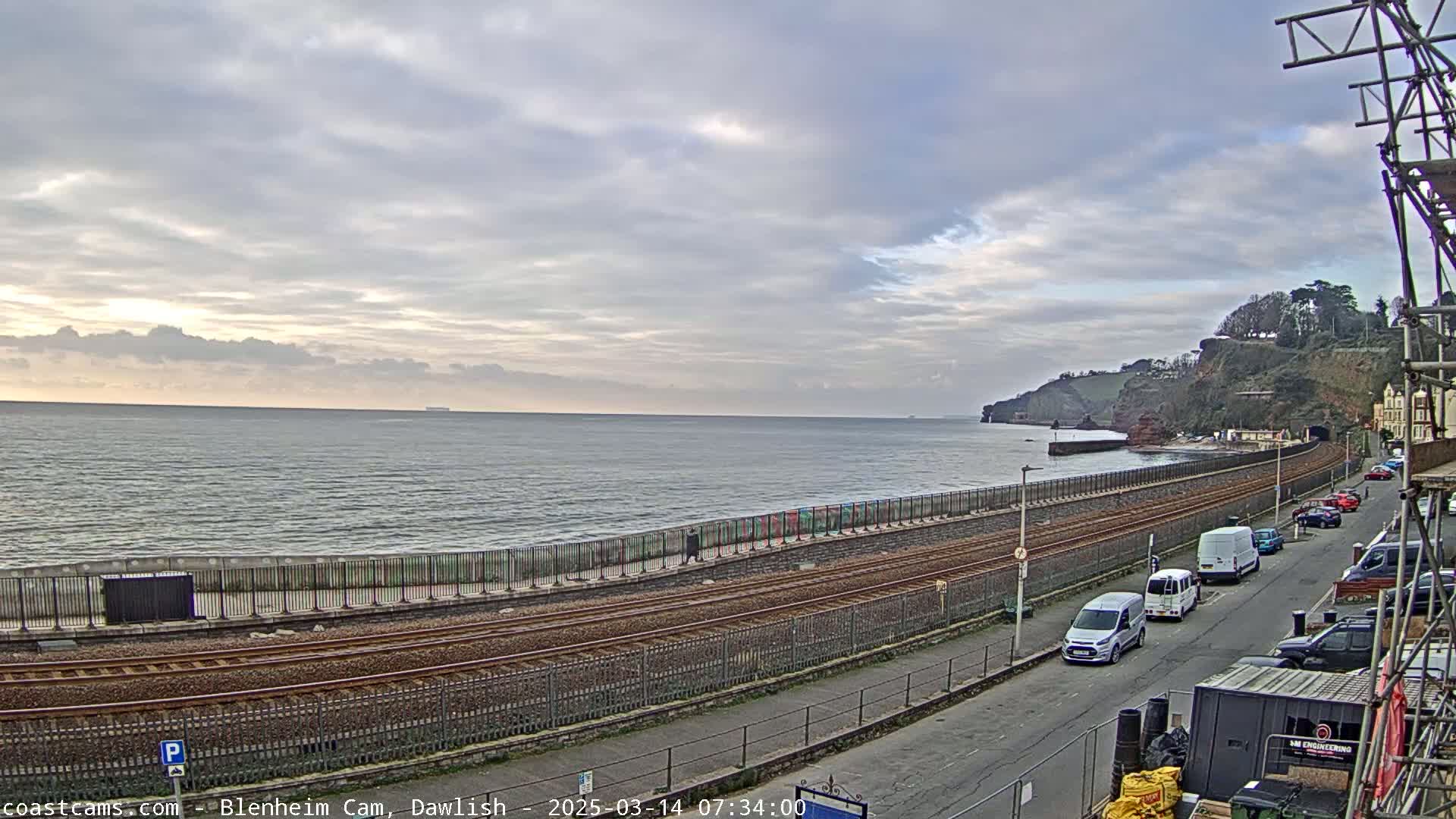 A coastal scene under a cloudy sky shows a train track running alongside a seawall, with several vehicles parked on a road beyond.