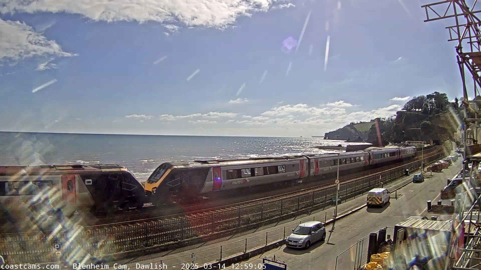 A long passenger train travels along a coastal railway line on a partly sunny day with a moderately wavy sea visible in the background.