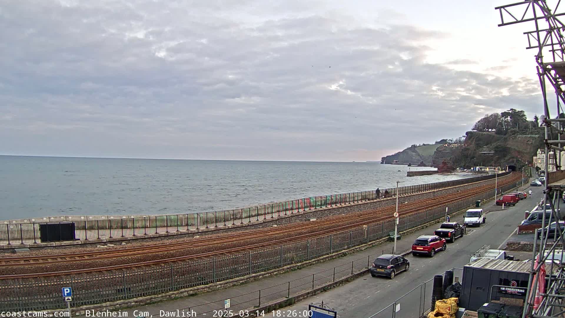 A coastal scene under an overcast sky shows a railway track running alongside a sea wall, with cars parked on a road beside it, and a calm sea in the background.
