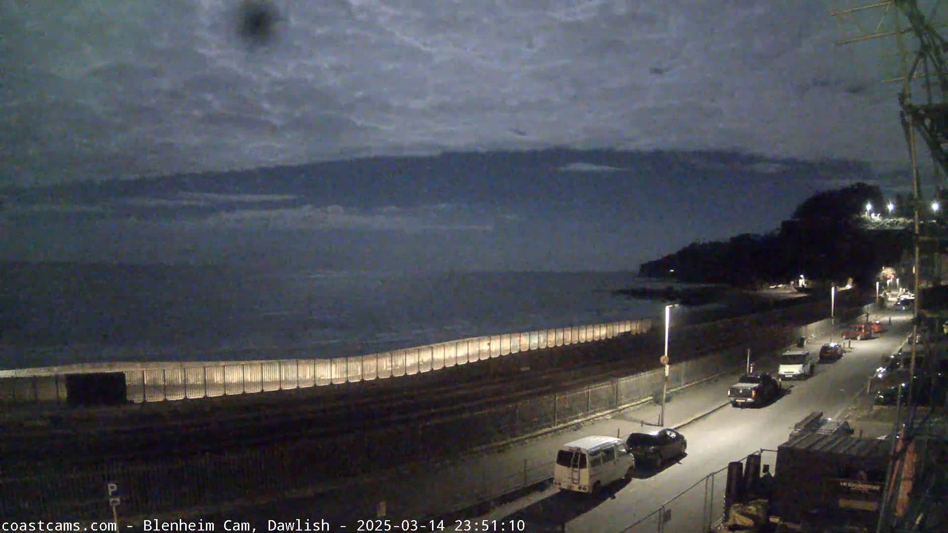 A nighttime coastal scene shows a dark ocean under a cloudy sky, with a road lined with parked cars and a seawall illuminated along the waterfront.