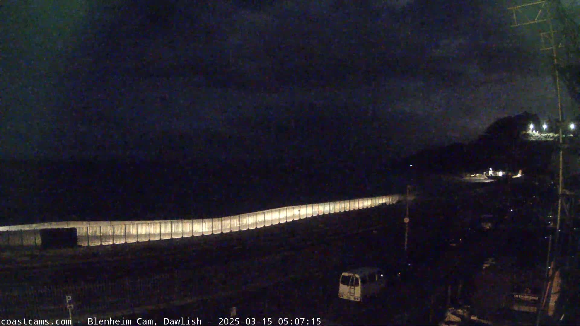 A long, illuminated seawall stretches across a dark, nighttime beach under a cloudy sky.