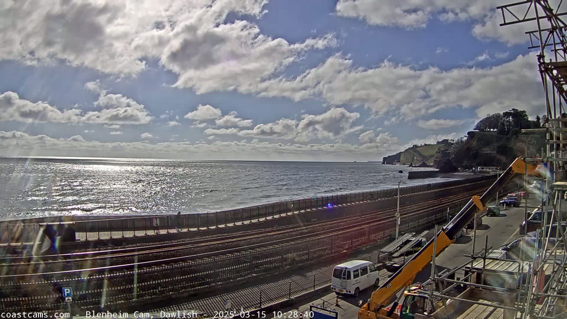 A partly sunny day shows a coastline with a train track running alongside the ocean, construction equipment present nearby.