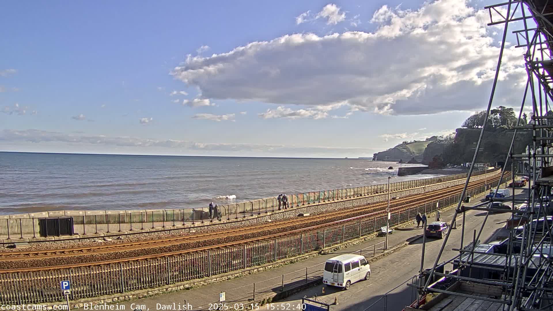 A partly cloudy day shows a coastal scene with a railway track running alongside a sea wall, a road, and parked cars, near a rocky cliffside.