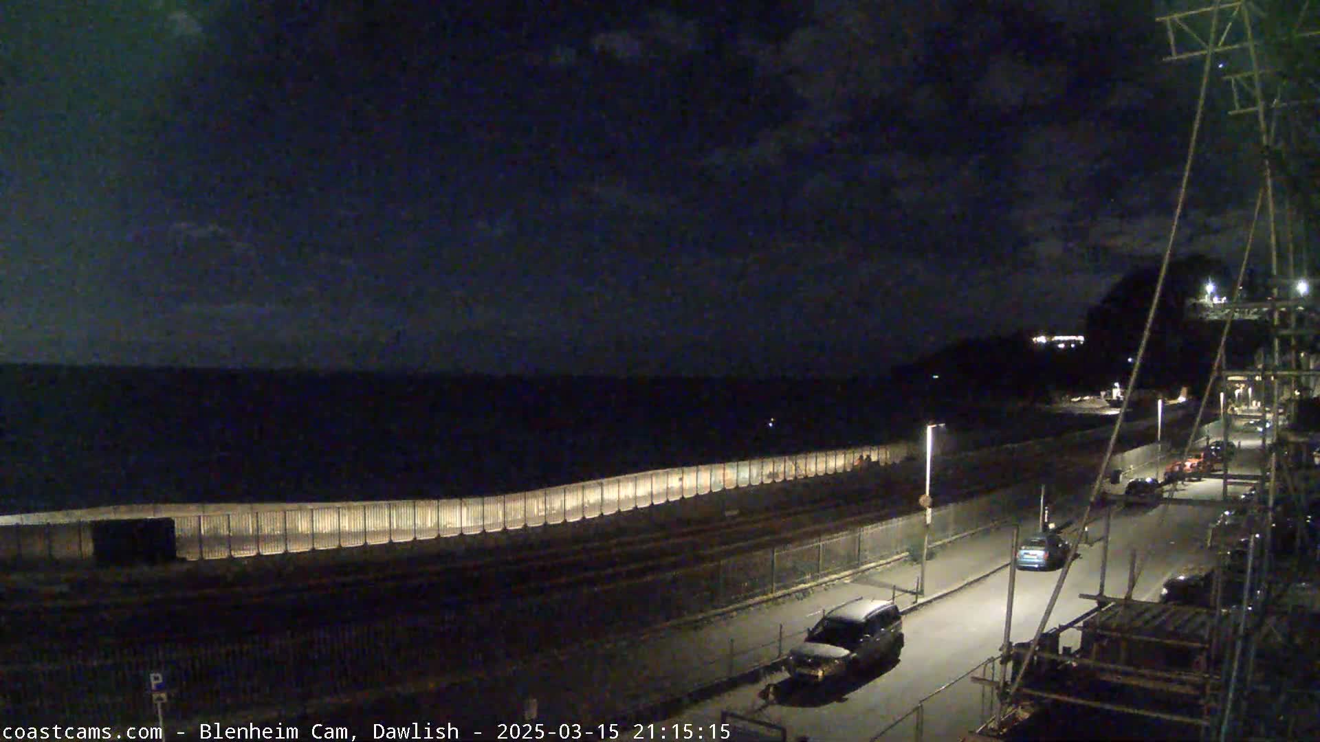 A nighttime coastal scene shows a dark sea, a long illuminated seawall, a road with a few parked and moving cars, and some construction scaffolding.