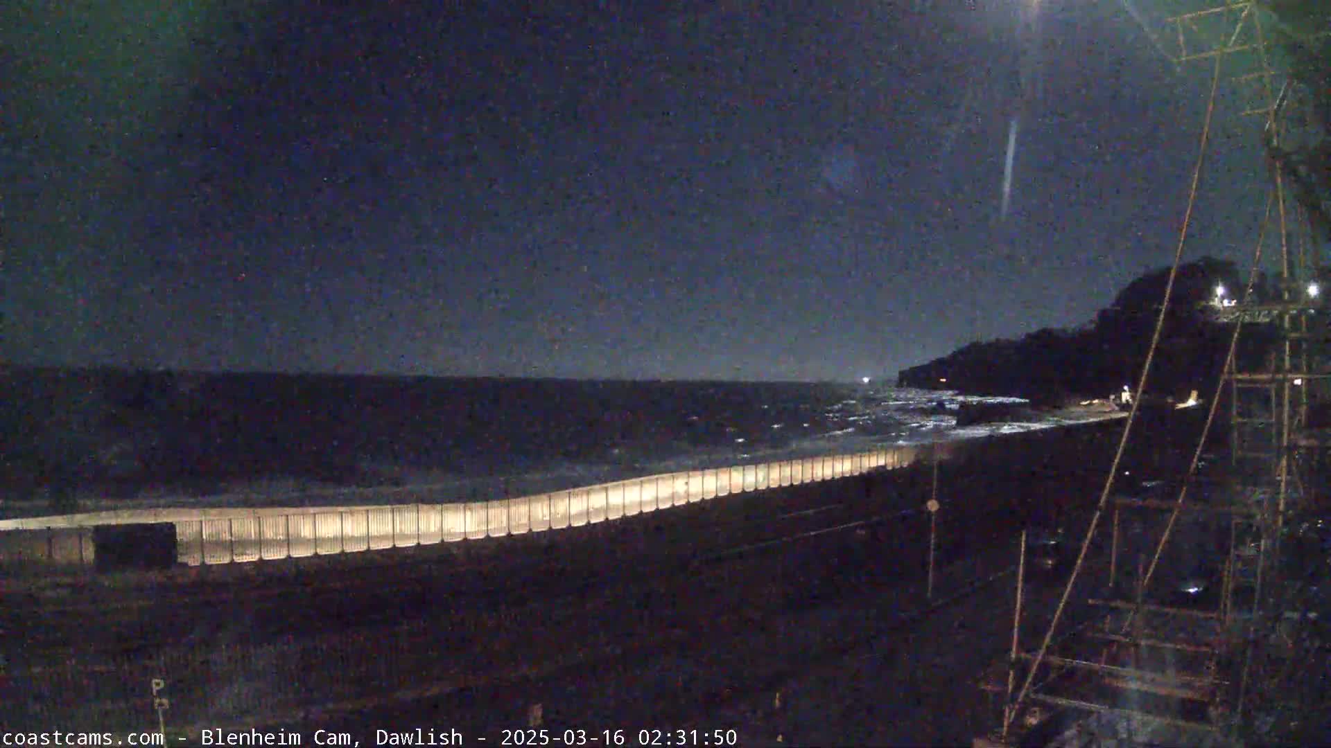 A nighttime view of a dark, wavy ocean meeting a shoreline protected by a long, illuminated seawall under a dark, clear sky.