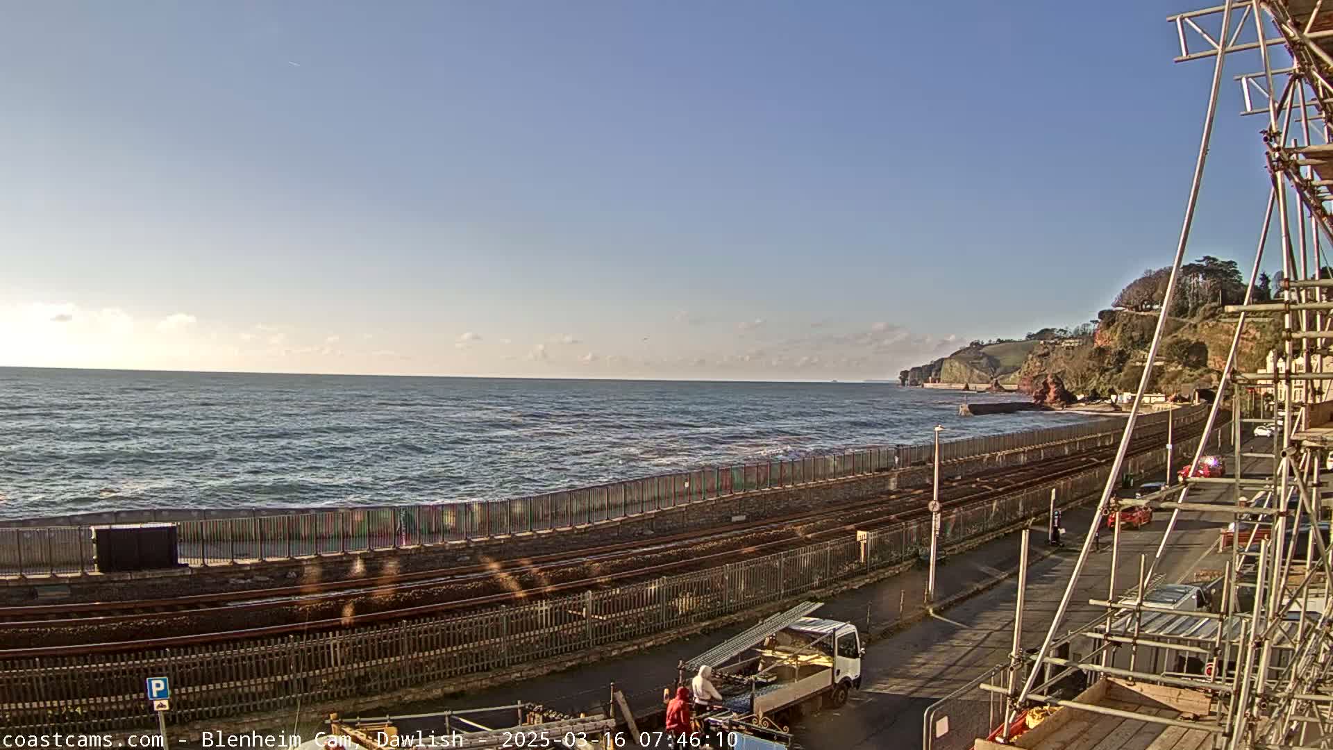 A coastal scene on a partly sunny day shows a railway line running alongside a sea wall, with a road and construction scaffolding visible in the foreground, and calm ocean waves breaking gently against the shore.