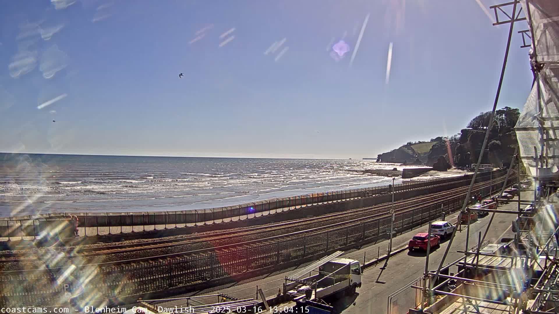 A sunny day at the beach shows a calm ocean with small waves, a railway track running parallel to the shore, cars parked along a road next to the track, and scaffolding in the foreground.