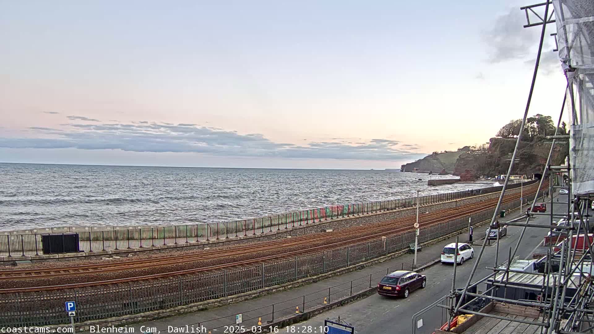 A slightly choppy sea stretches to a rocky coastline under a mostly cloudy, pale sky, with a railway line and road running alongside it, and several cars parked and driving along the road.
