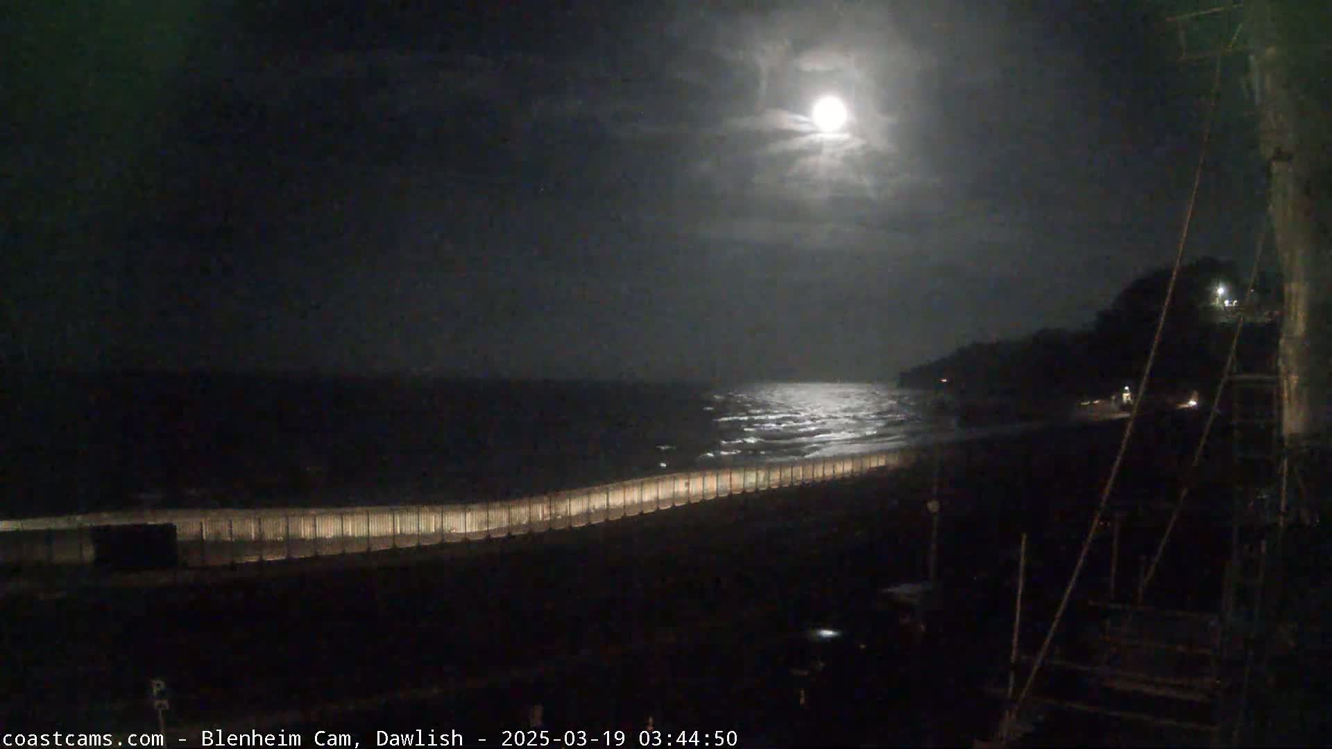 A nighttime view of a moonlit ocean with waves gently lapping against a seawall under a partly cloudy sky.