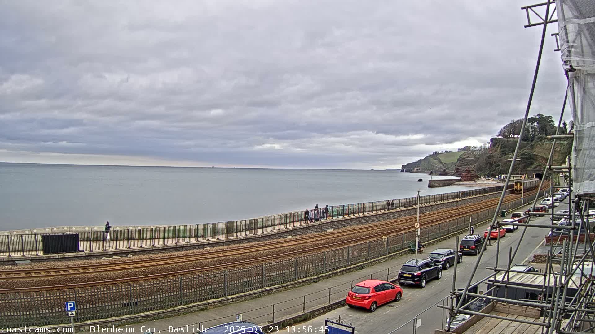 A coastal scene under a cloudy sky shows a train track running alongside a seawall with people walking on the wall and cars parked along a street beside it.