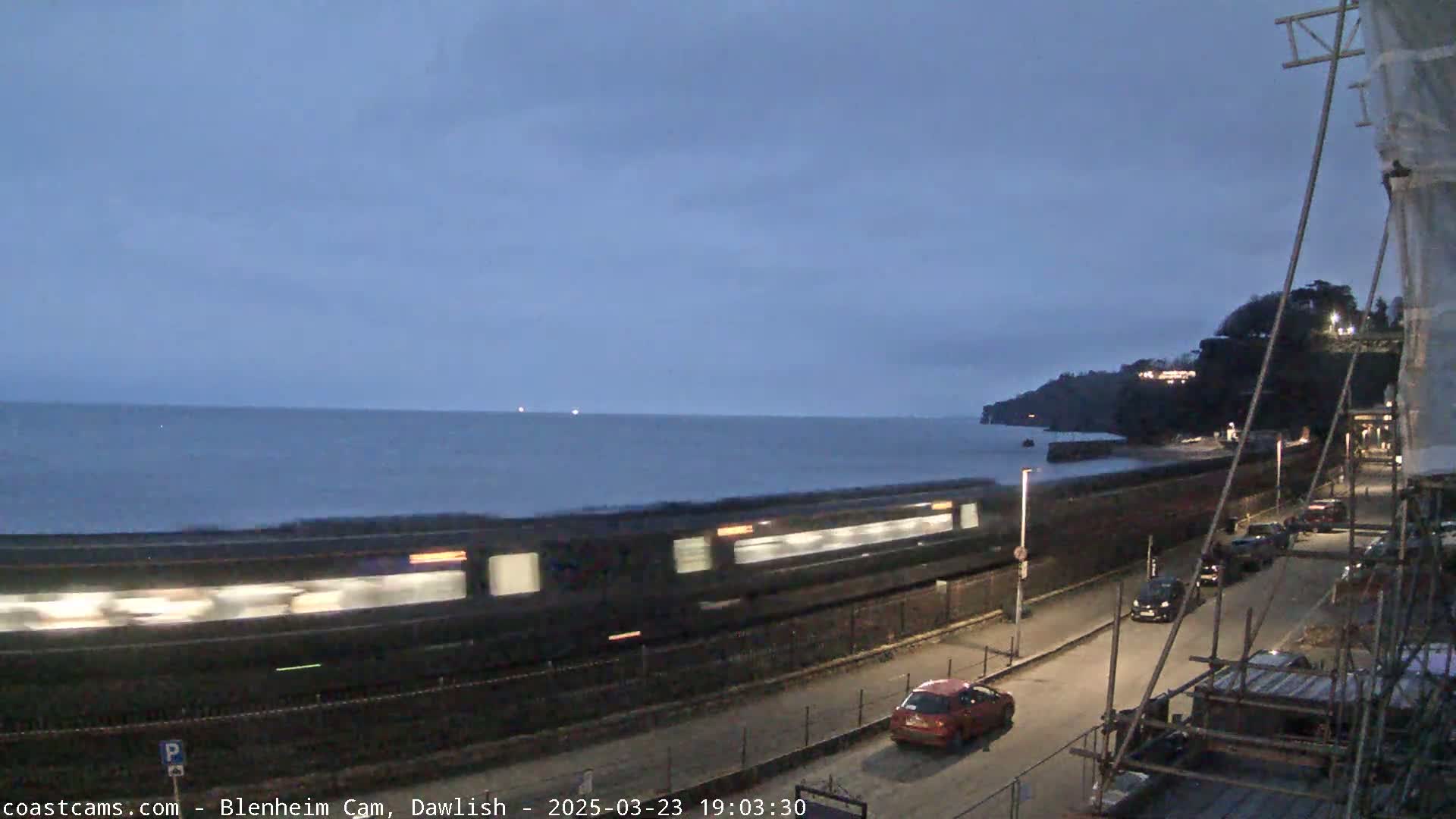 A long train passes by a seaside road at dusk, with a few cars parked along the road and a calm sea in the background.