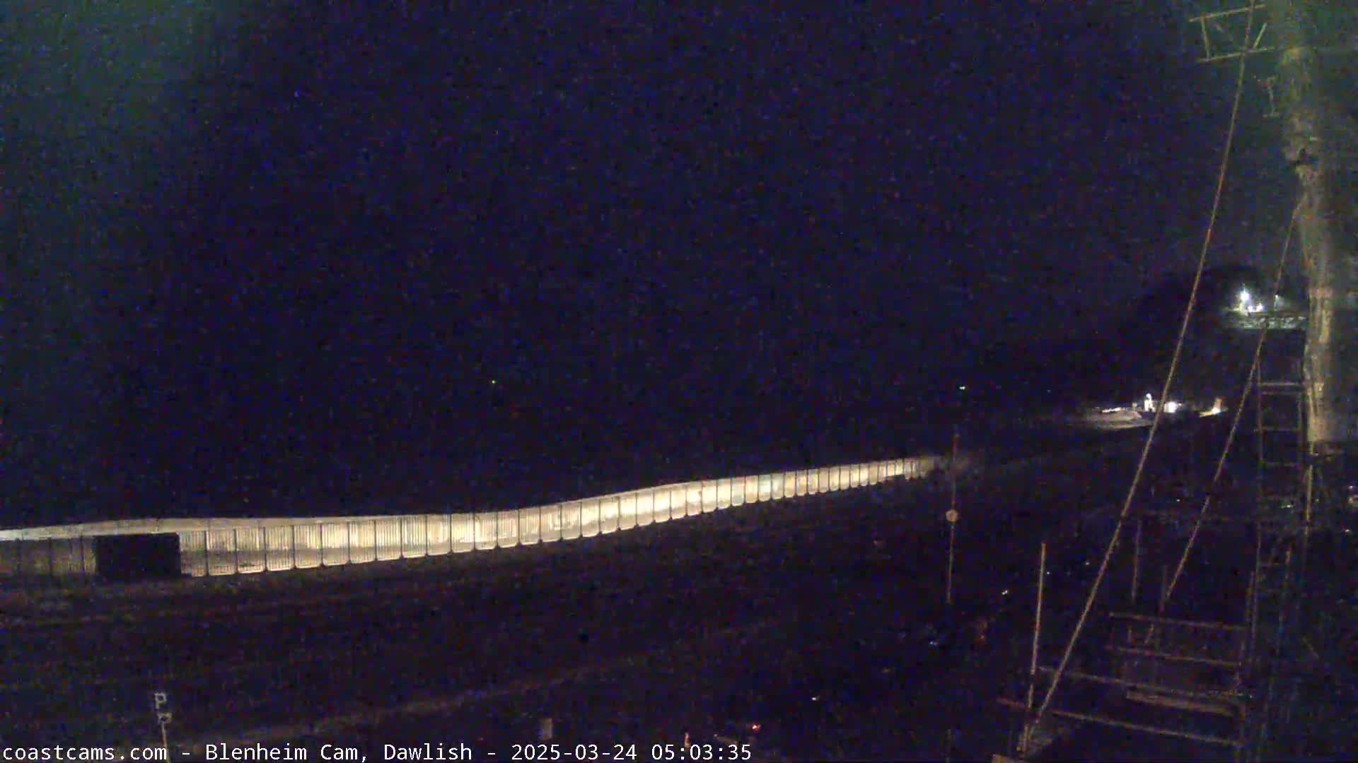 A long, illuminated barrier stretches across a dark, nighttime scene, with some indistinct objects visible in the distance.