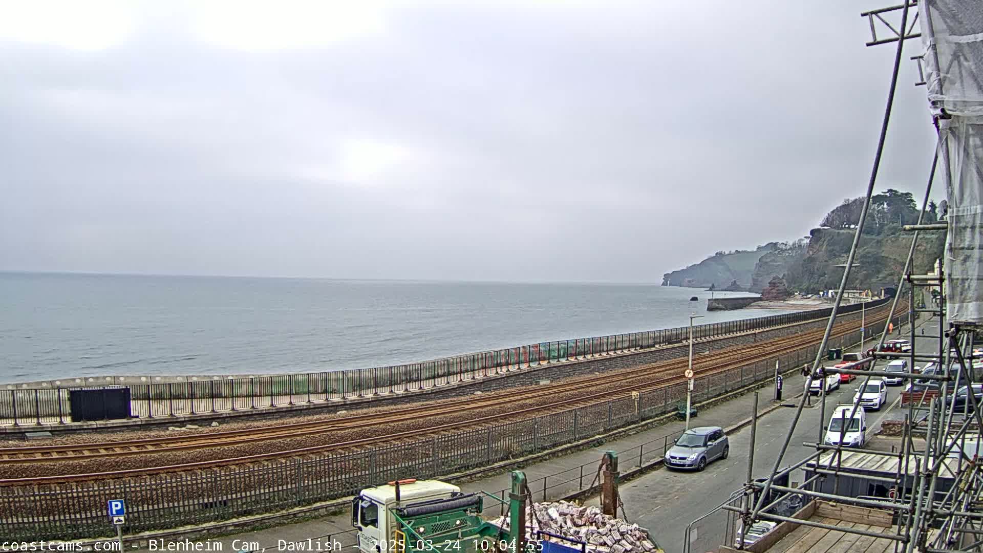 A coastal scene on an overcast day shows a railway track running alongside a sea wall, with a road and parked cars beyond, all leading to a beach and cliffs in the distance.