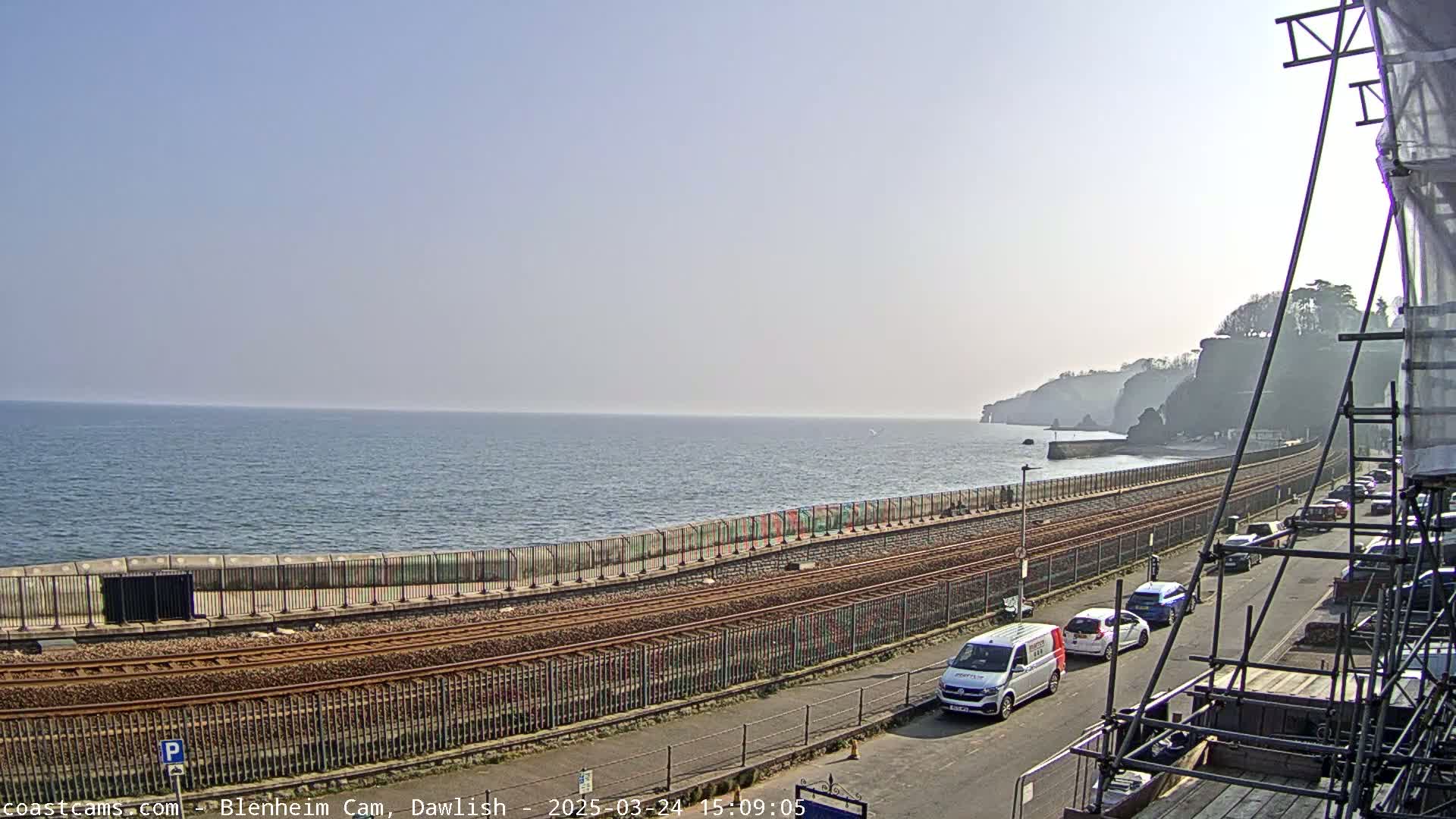 A coastal railway line runs alongside a sea wall, with several cars parked on a street next to scaffolding, under a clear, bright sky.