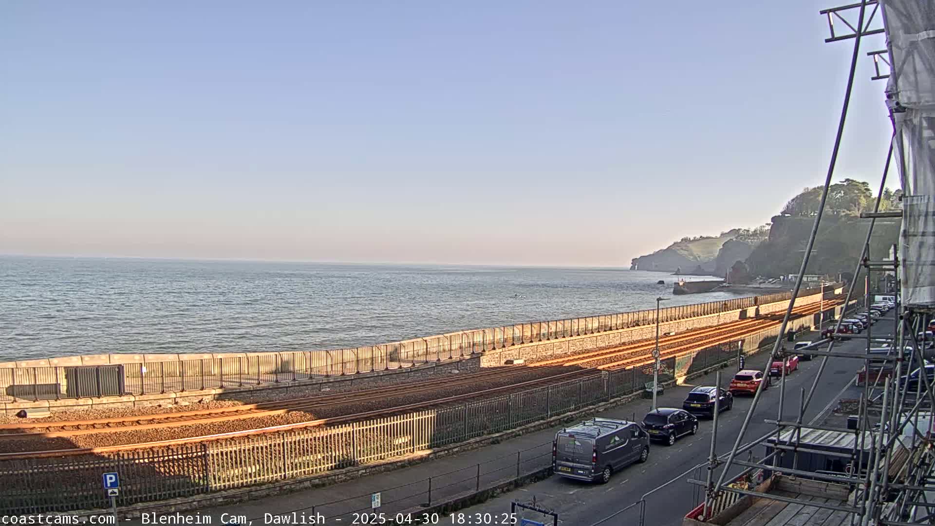 A calm sea stretches to a coastline with railway tracks, a road with parked cars, and cliffs in the distance under a clear sky.