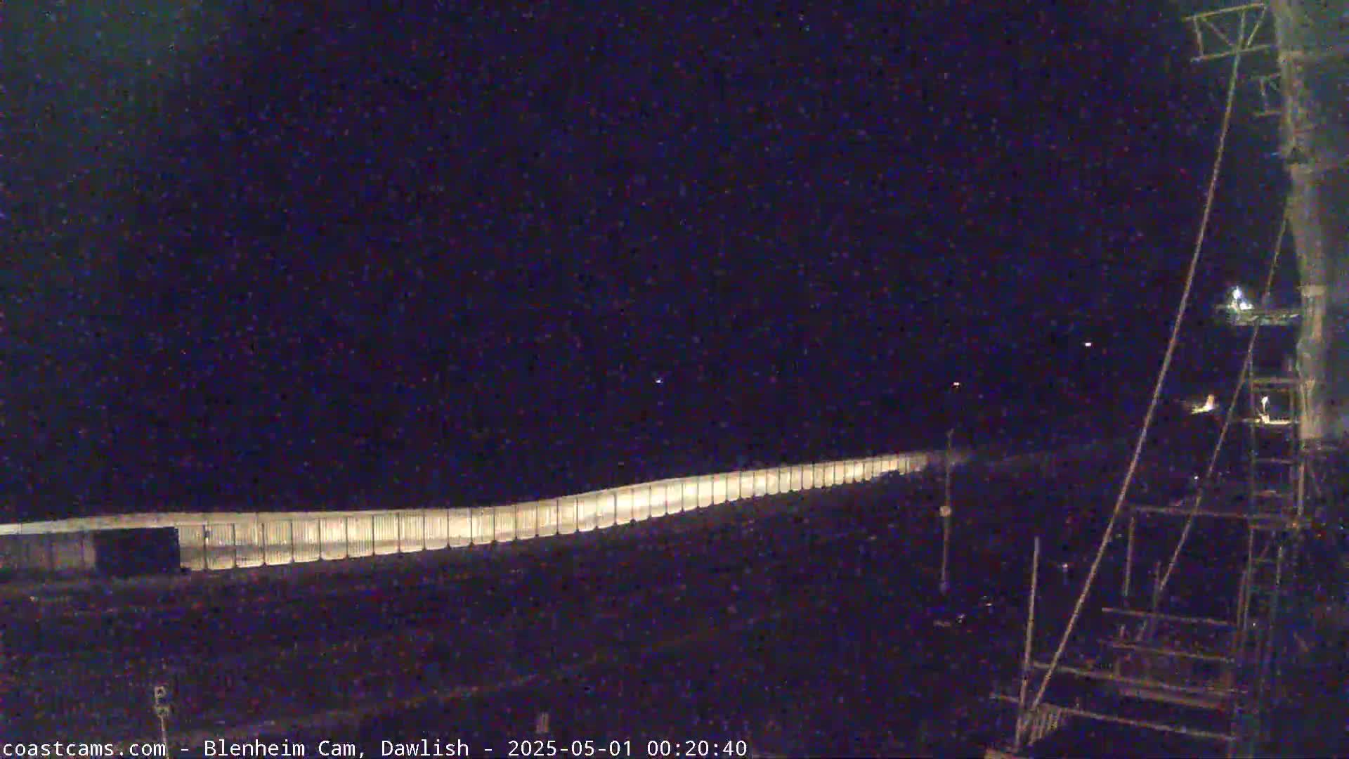 A long, illuminated barrier stretches across a dark, nighttime scene, with a tall metal structure in the lower right corner.