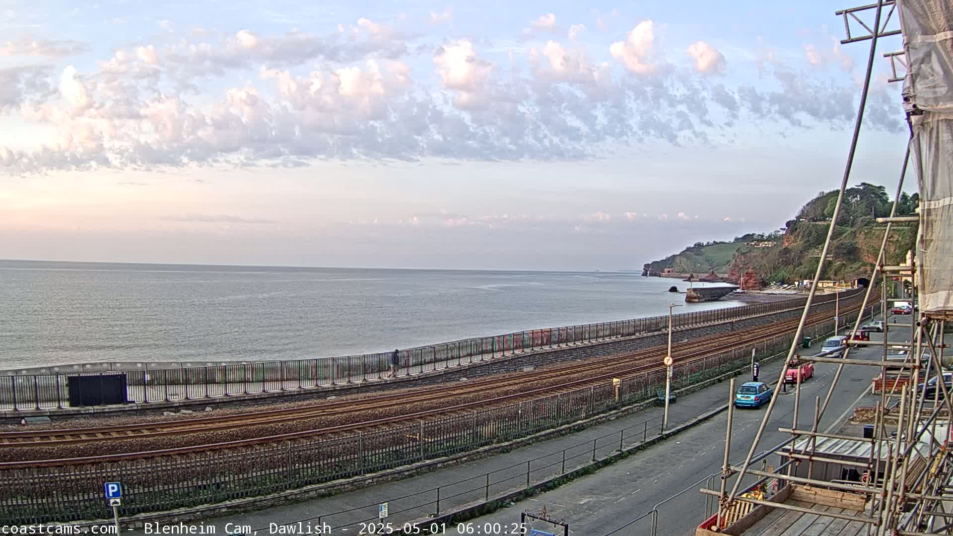 A partly cloudy sky overlooks a calm sea, railroad tracks, and a coastal road with several parked cars near construction scaffolding.