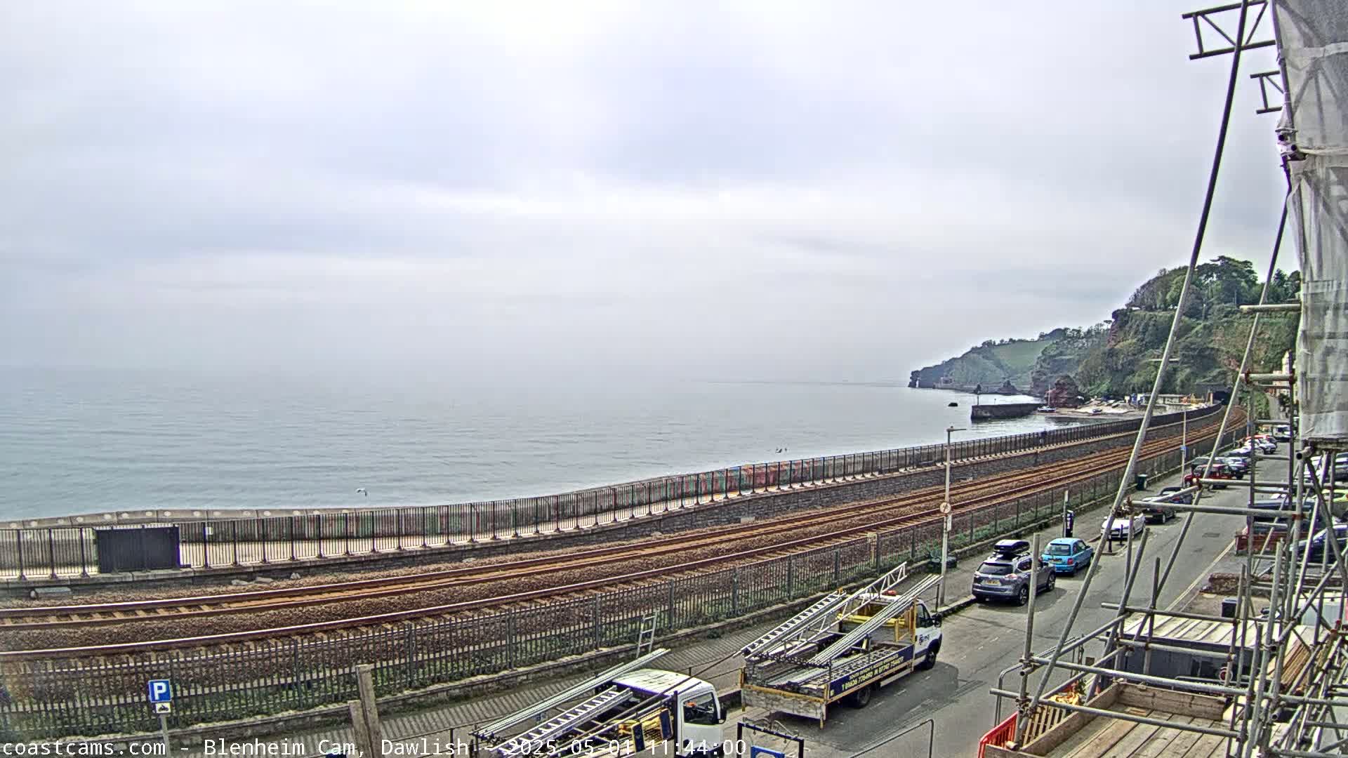 A coastal scene on an overcast day shows train tracks running alongside a seawall, with a road and parked cars visible beyond, and a partially obscured view of the ocean and a cliffside in the distance.