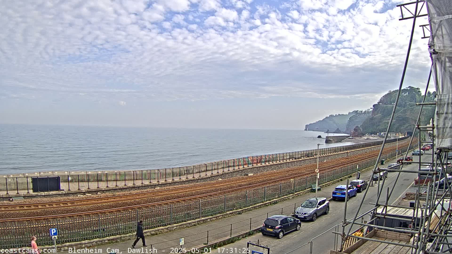 A seaside scene under a partly cloudy sky shows a train track running along a seawall, with cars parked along a street next to it, and a cliff in the distance.