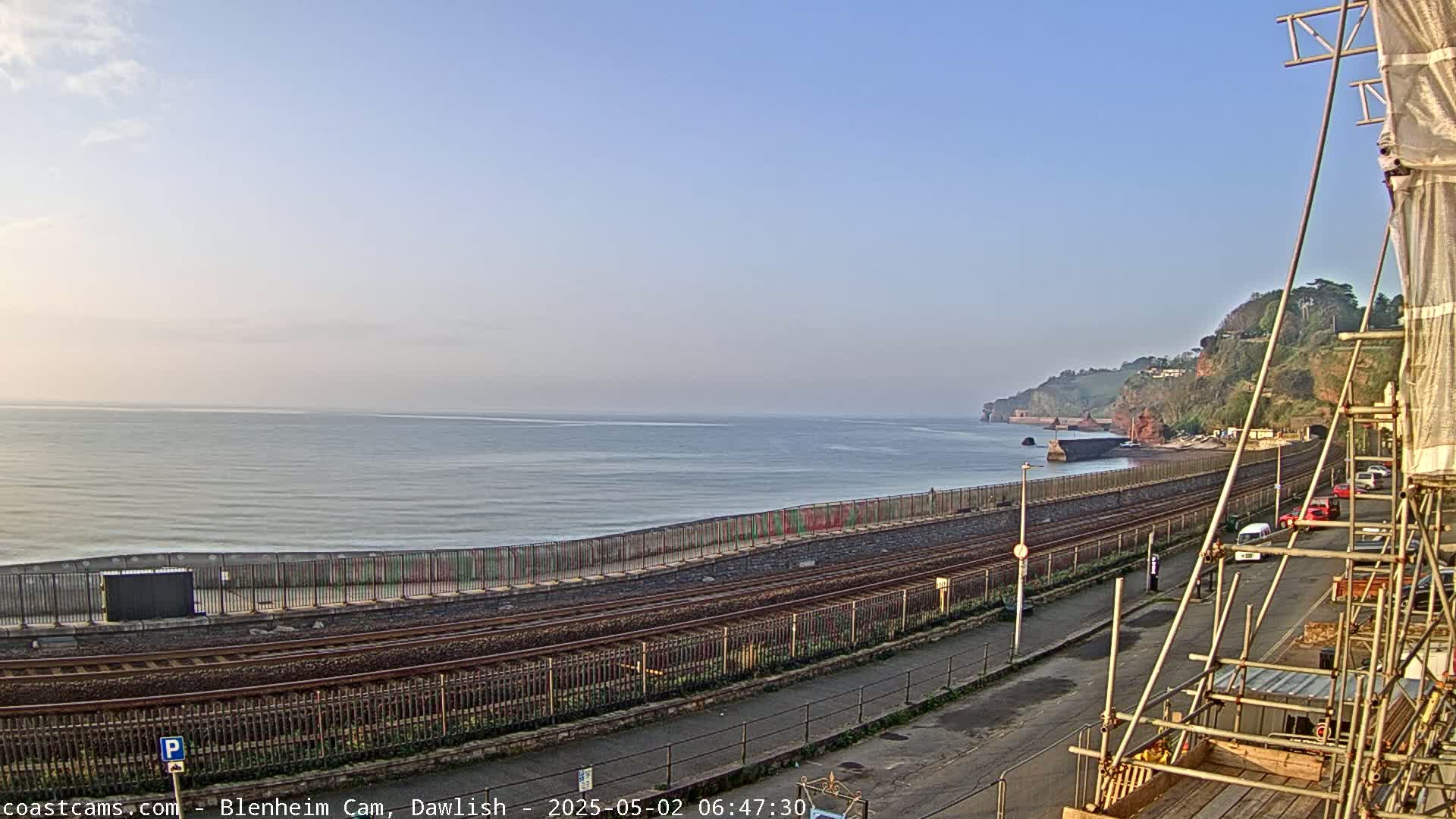 A calm ocean stretches to the horizon under a clear, light-blue sky, beside a railway line and seawall that runs alongside a road, past cliffs and some parked cars near construction scaffolding.