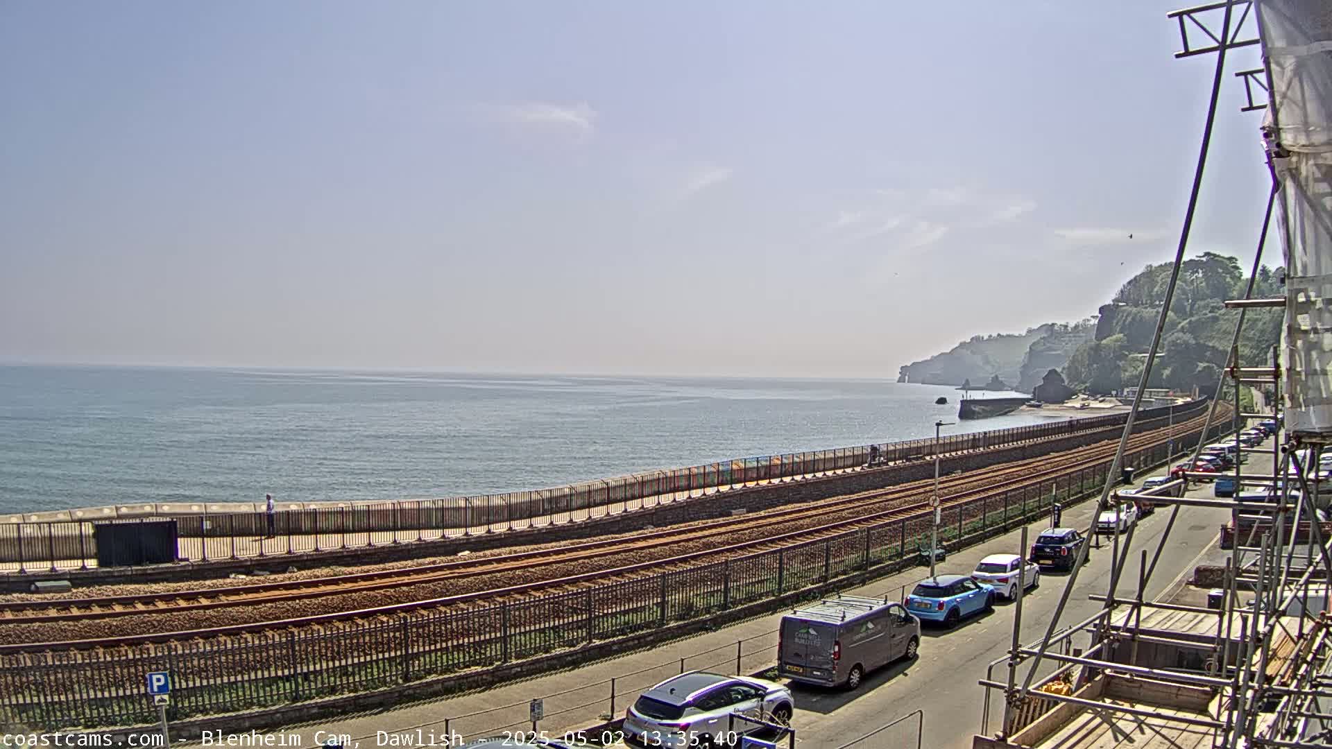 A seaside view on a sunny day shows a train track running along a coastal path, with cars parked along a nearby road and scaffolding in the foreground.