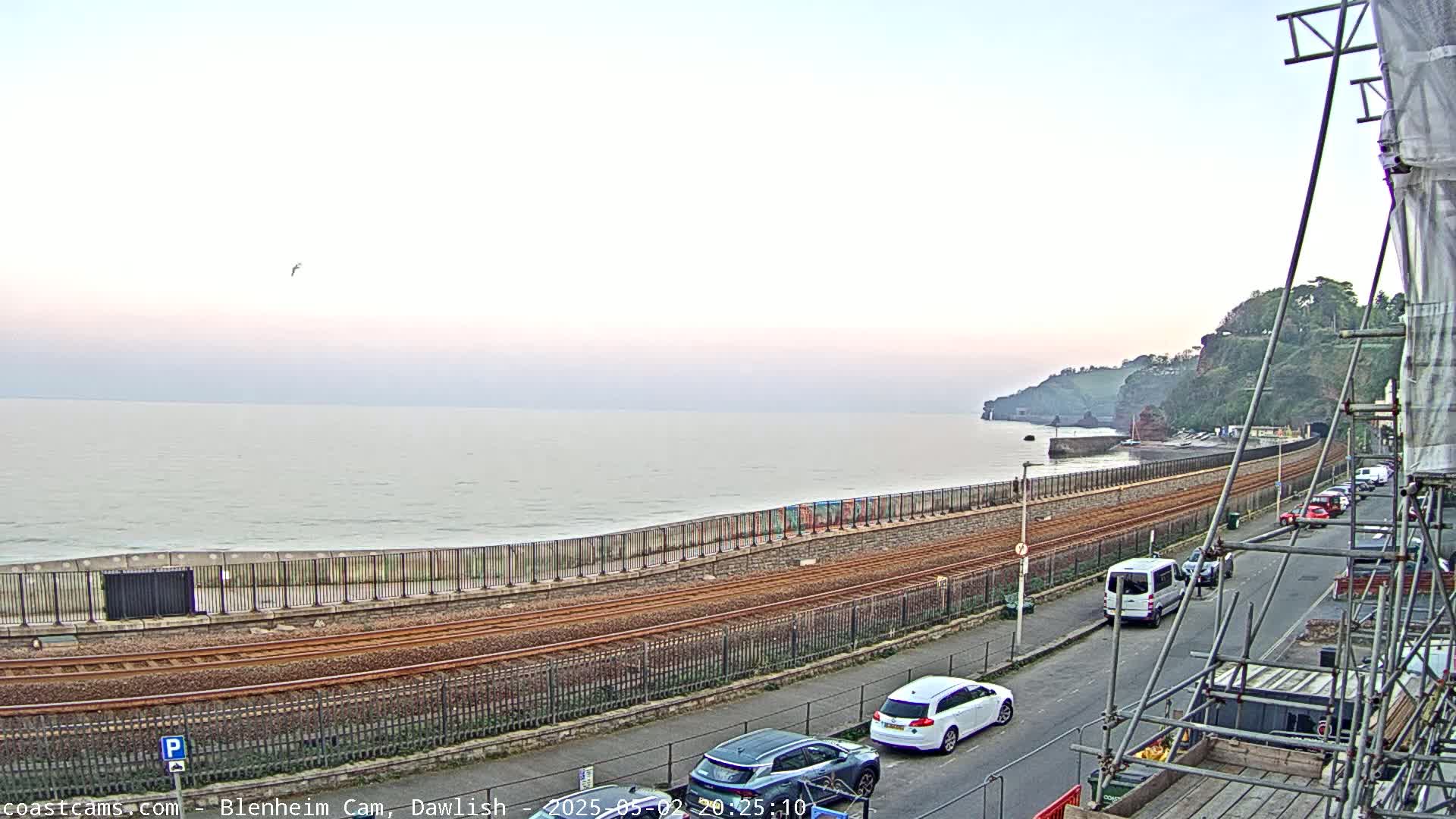 A calm sea stretches to a hazy horizon under a pale sky, beside a railway line bordering a road with parked cars and scaffolding.