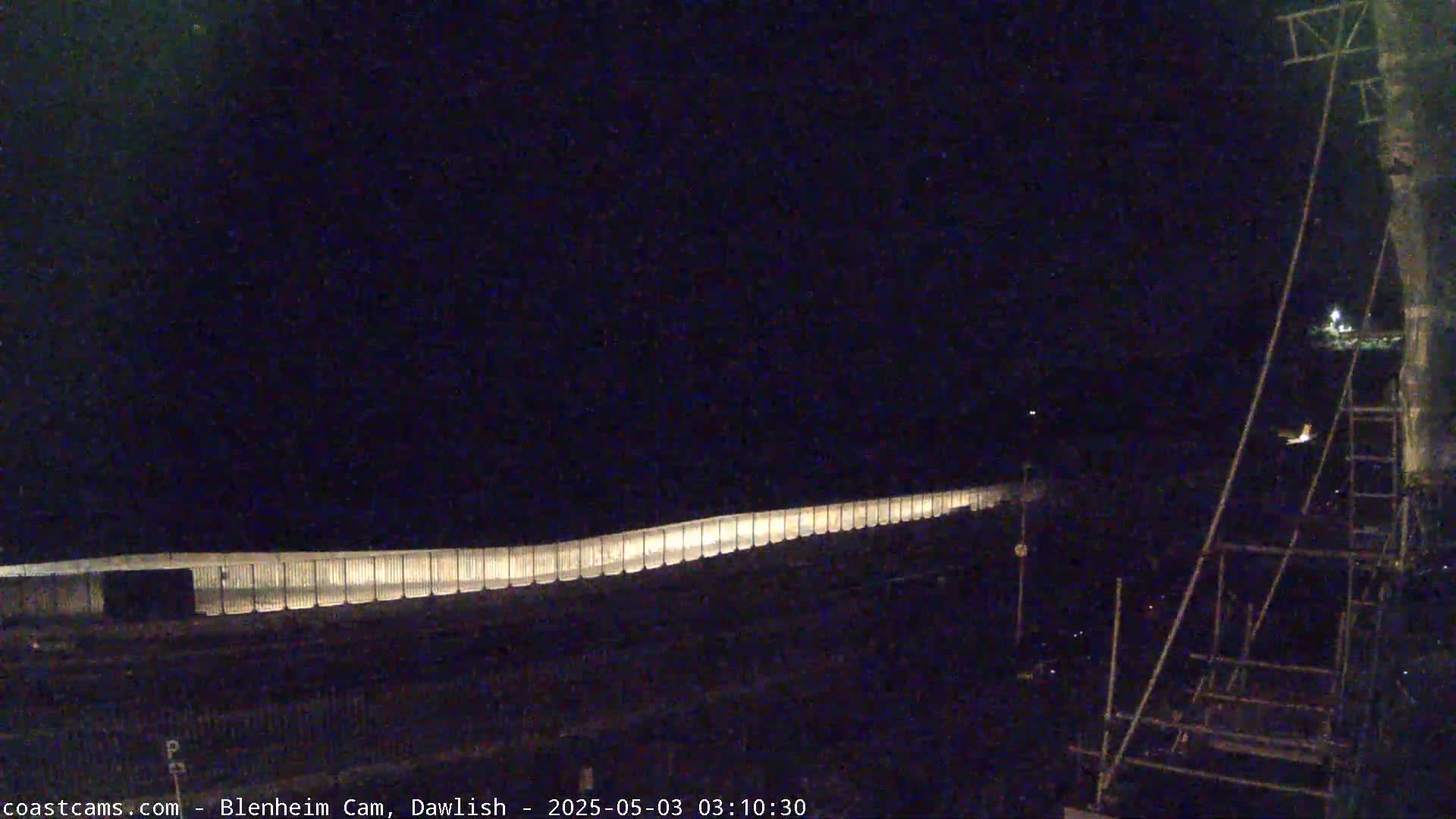 A nighttime long-exposure shot shows a lighted barrier stretching across the bottom of the frame, with scaffolding and other structures to the right.