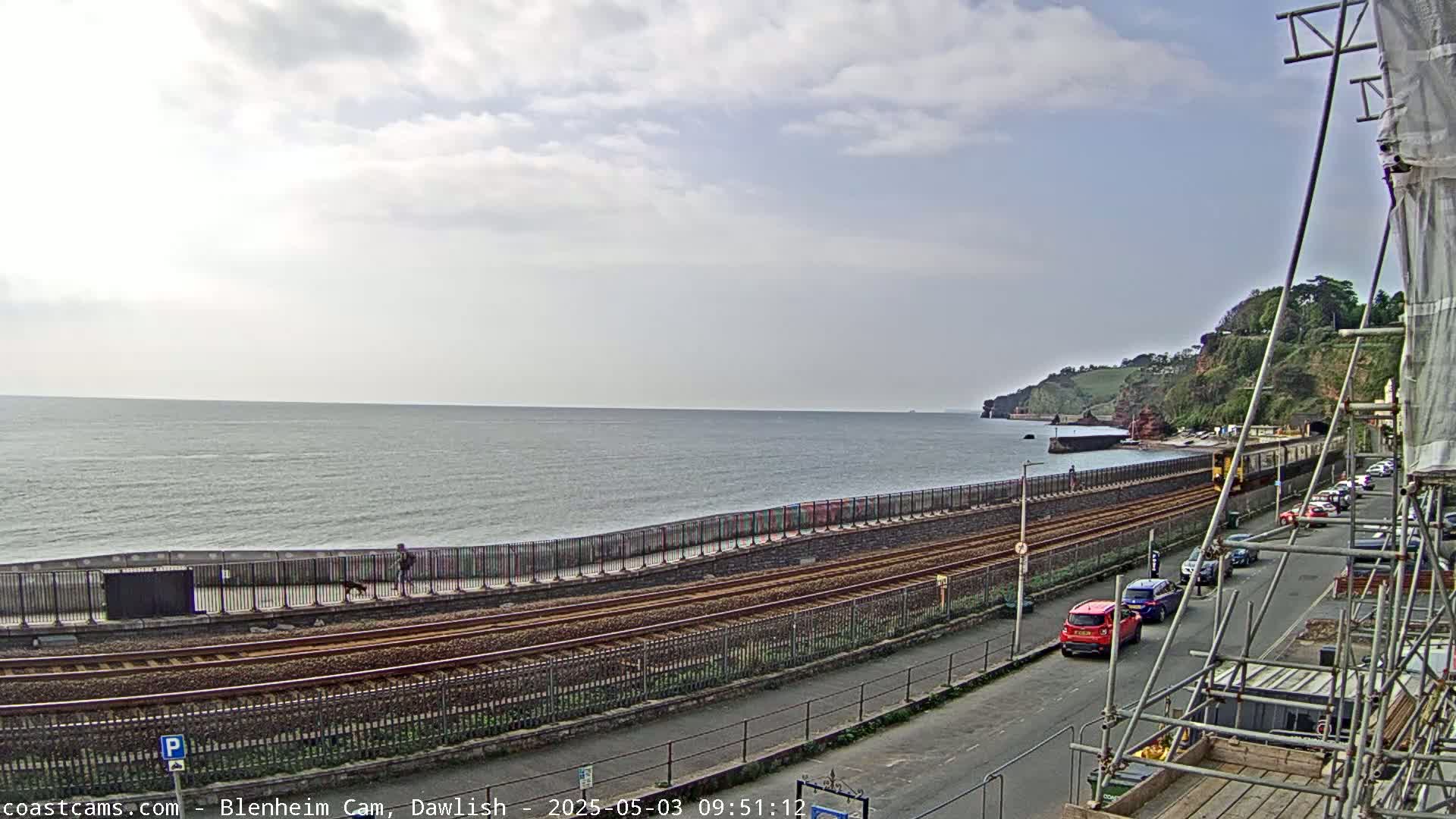 A mostly cloudy day shows a seaside town with train tracks running alongside a seawall, cars parked on a street, and a person walking a dog along the seawall.