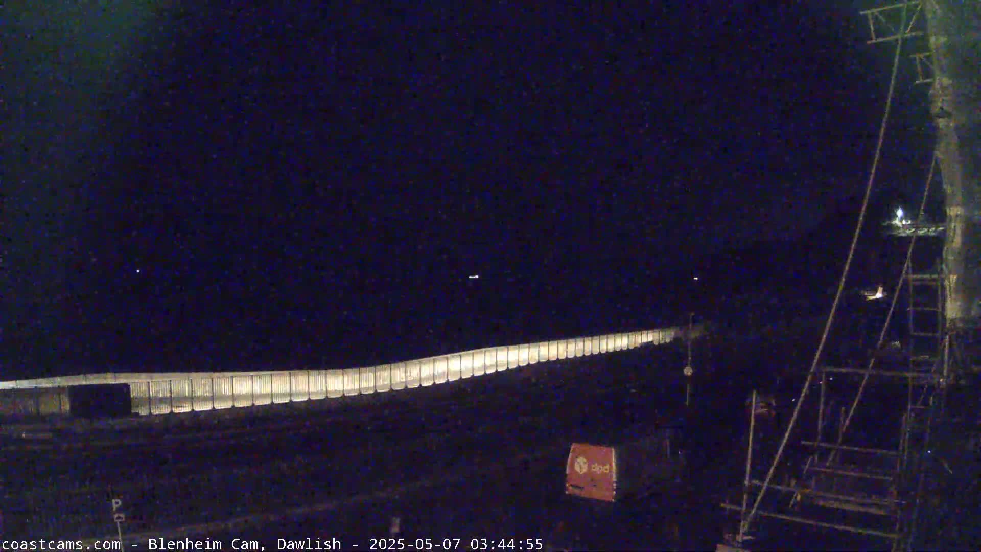 A long, illuminated barrier stretches across a dark, nighttime scene, with a portion of a structure and a vehicle visible in the lower right corner.