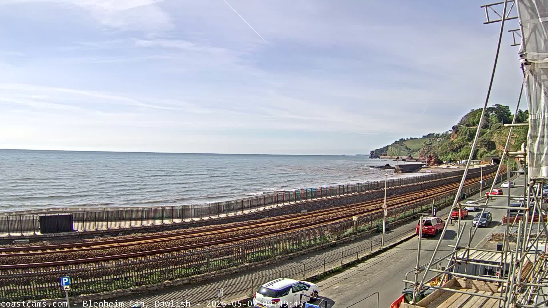 A calm ocean stretches to a coastline with railway tracks running alongside a road and parked cars, under a partly cloudy sky.