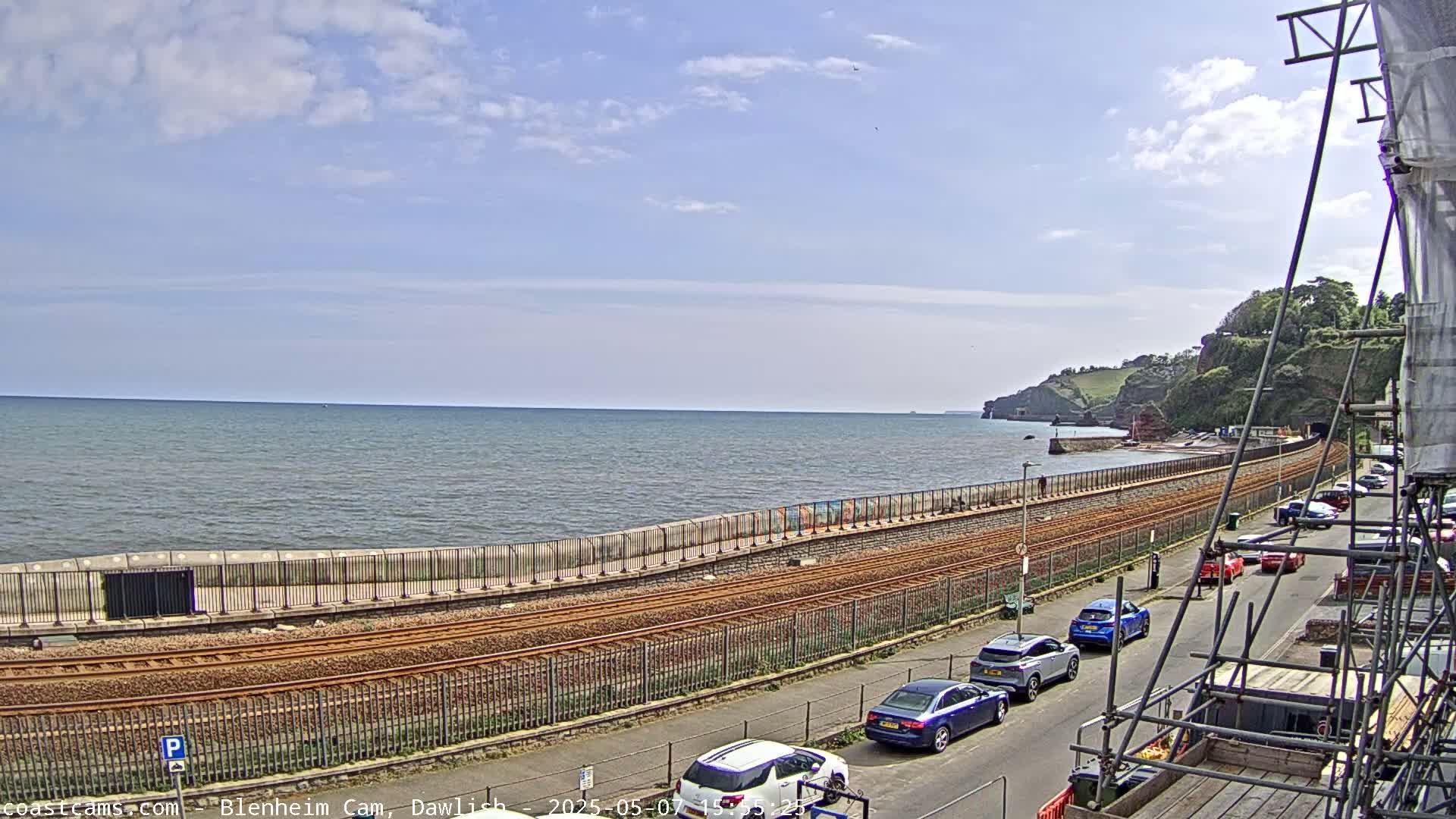 A partly cloudy day shows a calm ocean next to a seawall with train tracks and parked cars along a road.