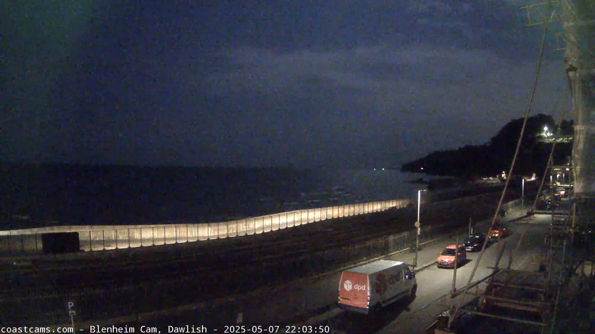 A dark, nighttime coastal scene shows a calm sea behind a lit seawall, with a few vehicles on a road next to it.