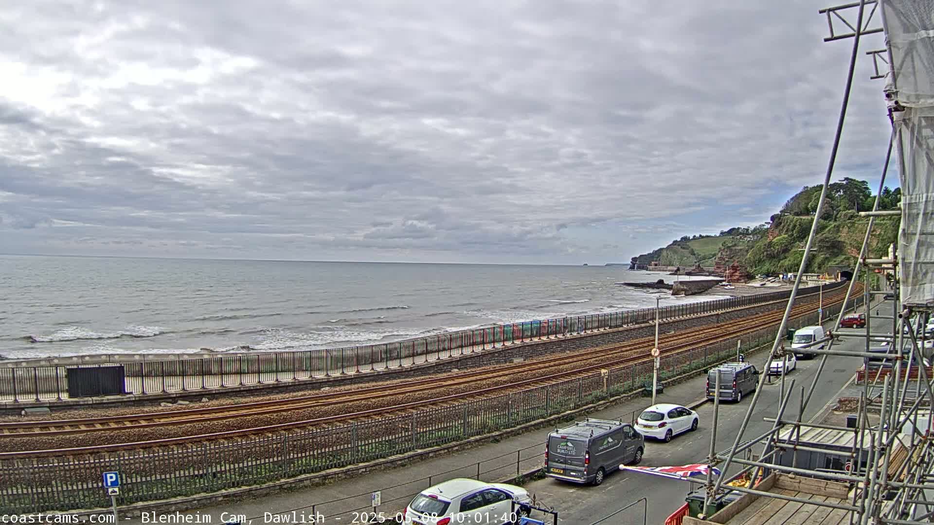 A coastal scene under a cloudy sky shows a train track running alongside a beach with a few cars parked along the road, and some construction scaffolding is visible.