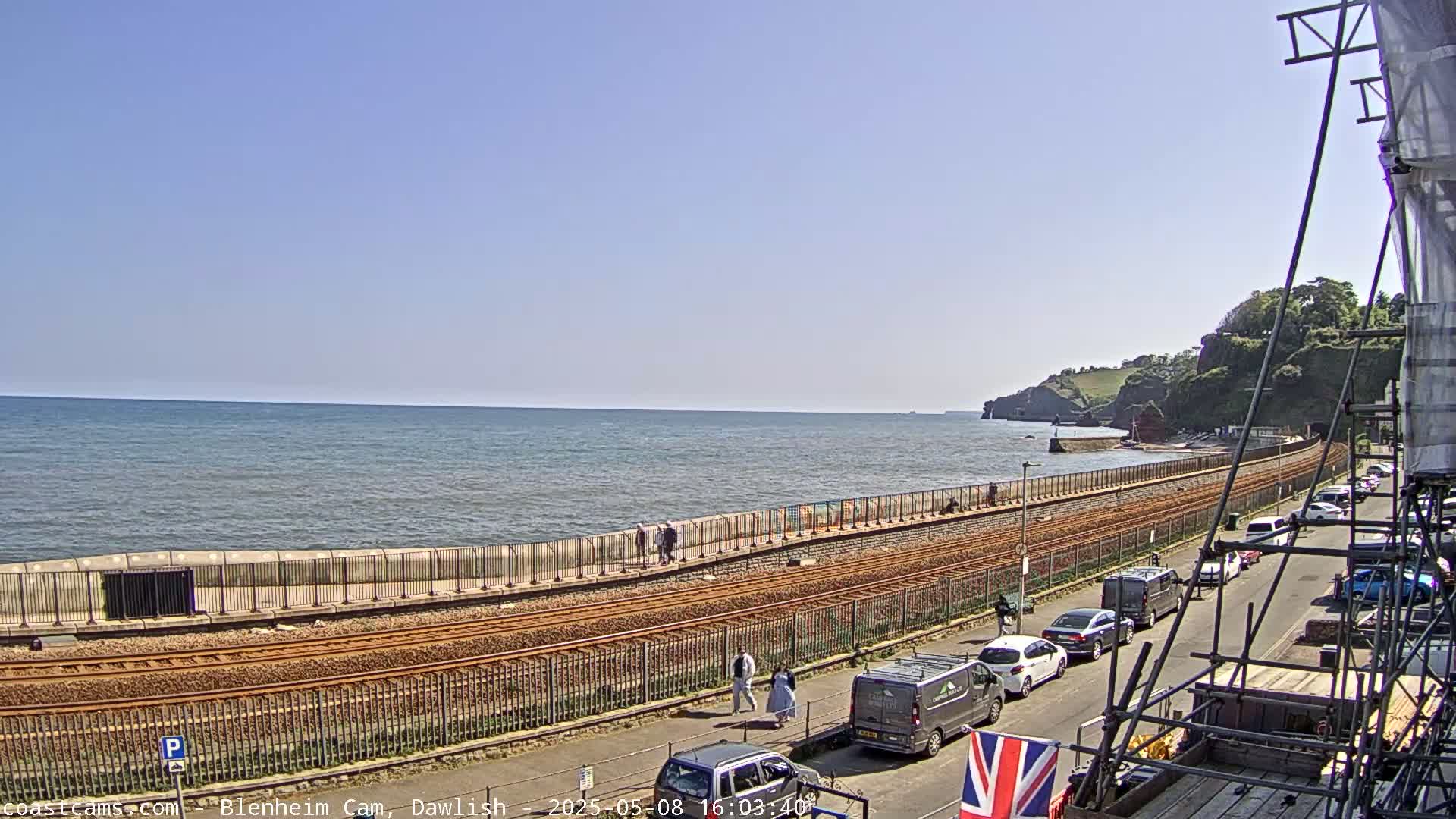 A seaside view on a sunny day shows a calm ocean, a seawall with a walkway and railway tracks, cars parked along a road, and people walking along the seawall.