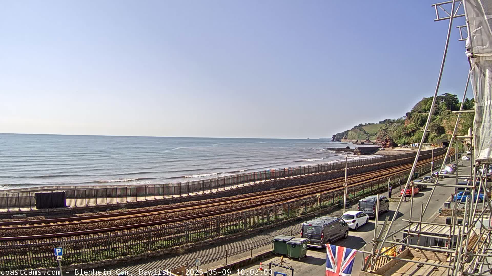 A seaside railway track runs alongside a calm ocean under a clear blue sky, with cars parked along a road next to it and construction scaffolding visible in the foreground.