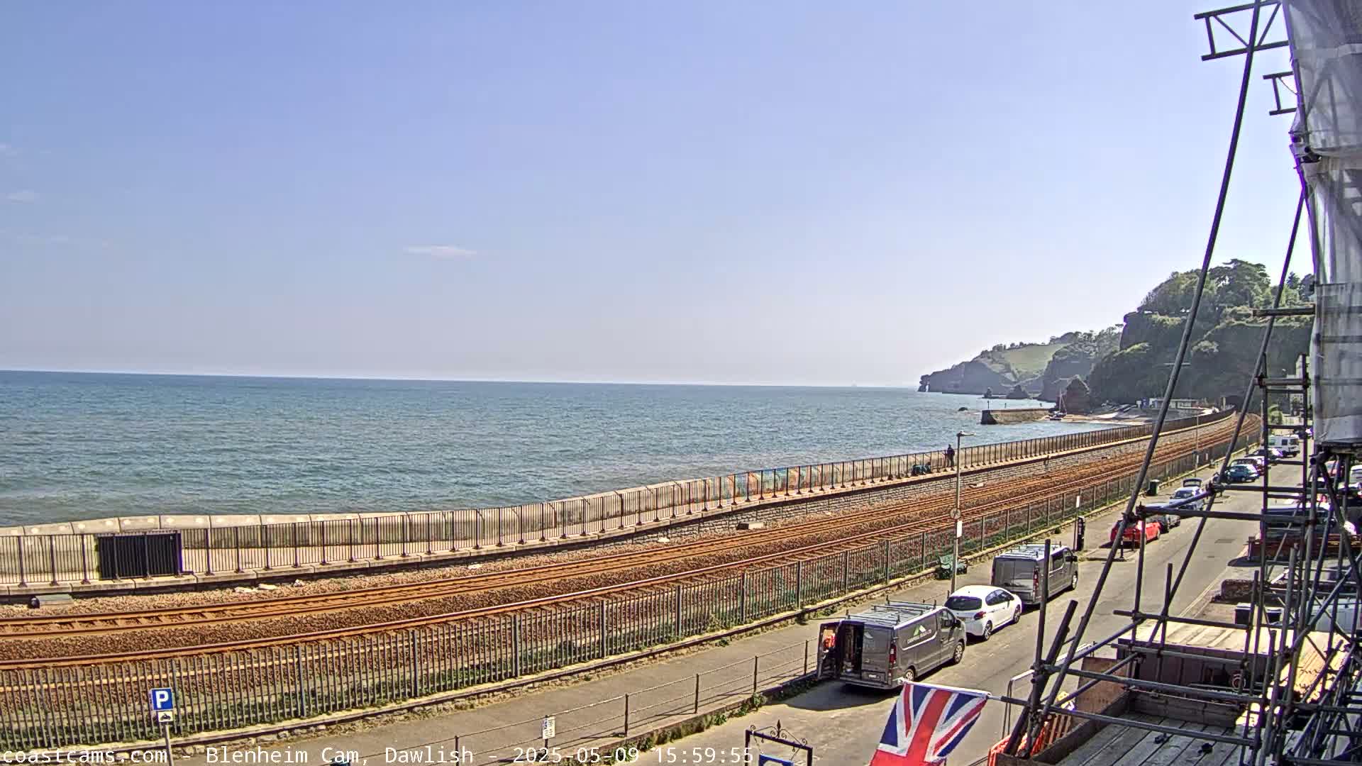 A coastal scene on a sunny day shows railway tracks running alongside a seawall, with cars parked on a road beyond the tracks, and cliffs visible in the distance.