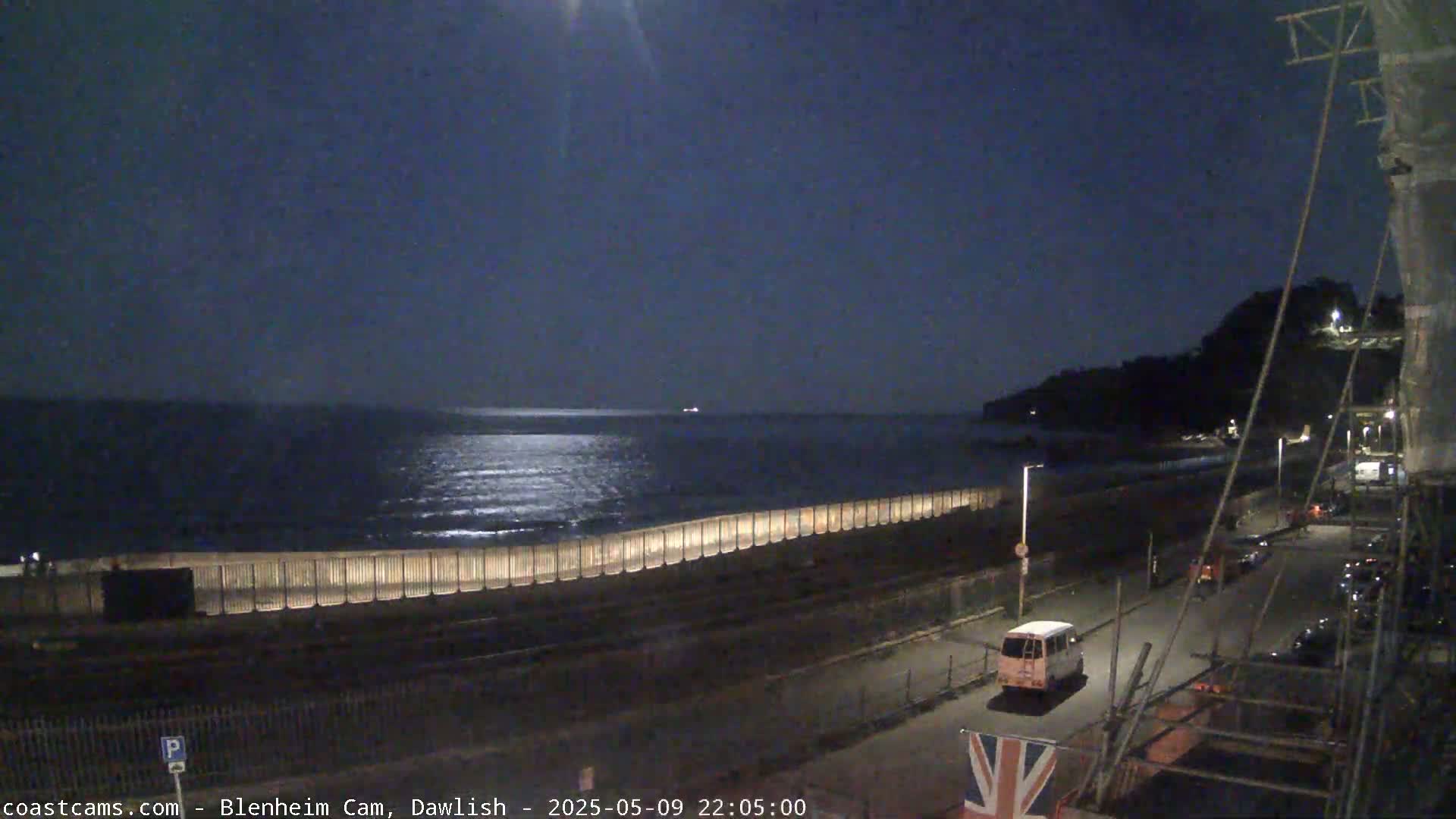 A moonlit nighttime coastal scene shows a calm sea, a seawall, a road with a few vehicles, and a hill in the background.