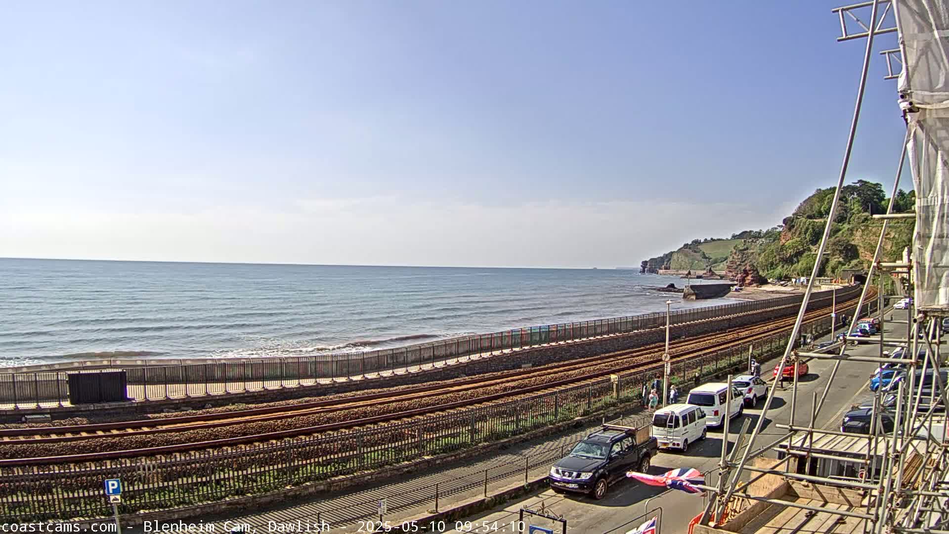 A seaside view shows a calm ocean, railway tracks running along a shoreline, parked cars on a street next to the tracks, and scaffolding in the foreground under a mostly sunny sky.