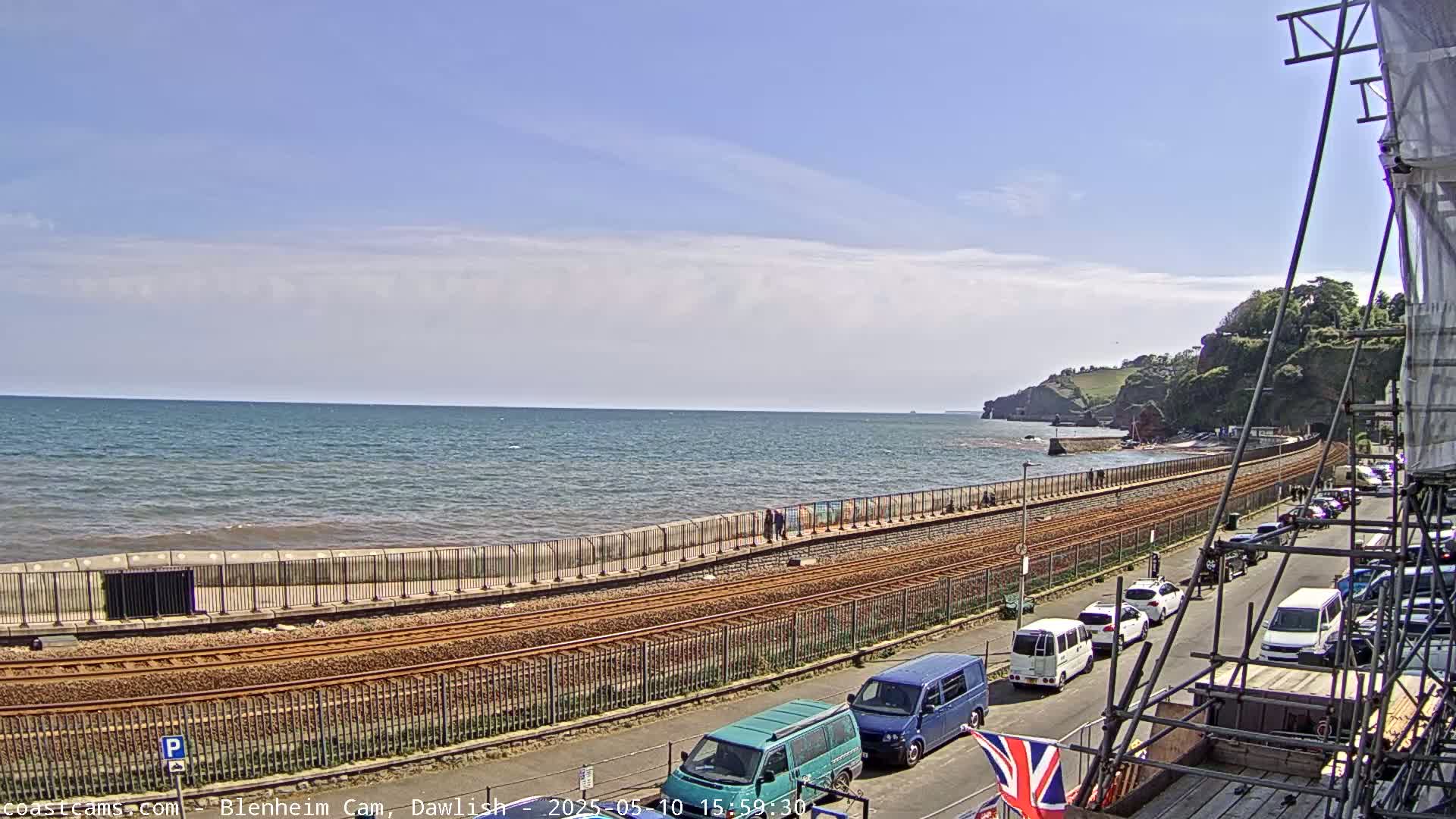 A mostly sunny day shows a seaside town with a train track running along a seawall, several cars parked along the road, and a partially obscured view of the ocean.