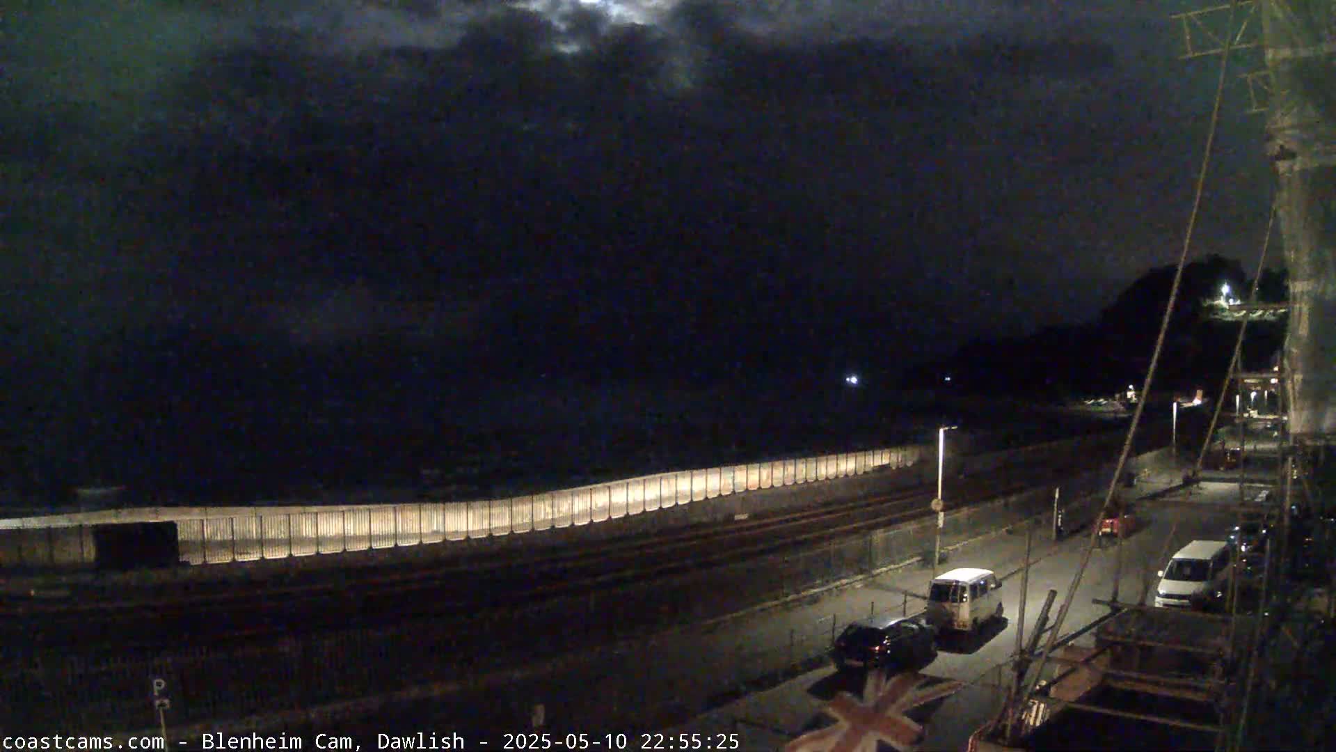 A nighttime coastal scene shows a dark, cloudy sky over a sea wall, a road with several cars parked along it, and a hill in the background.