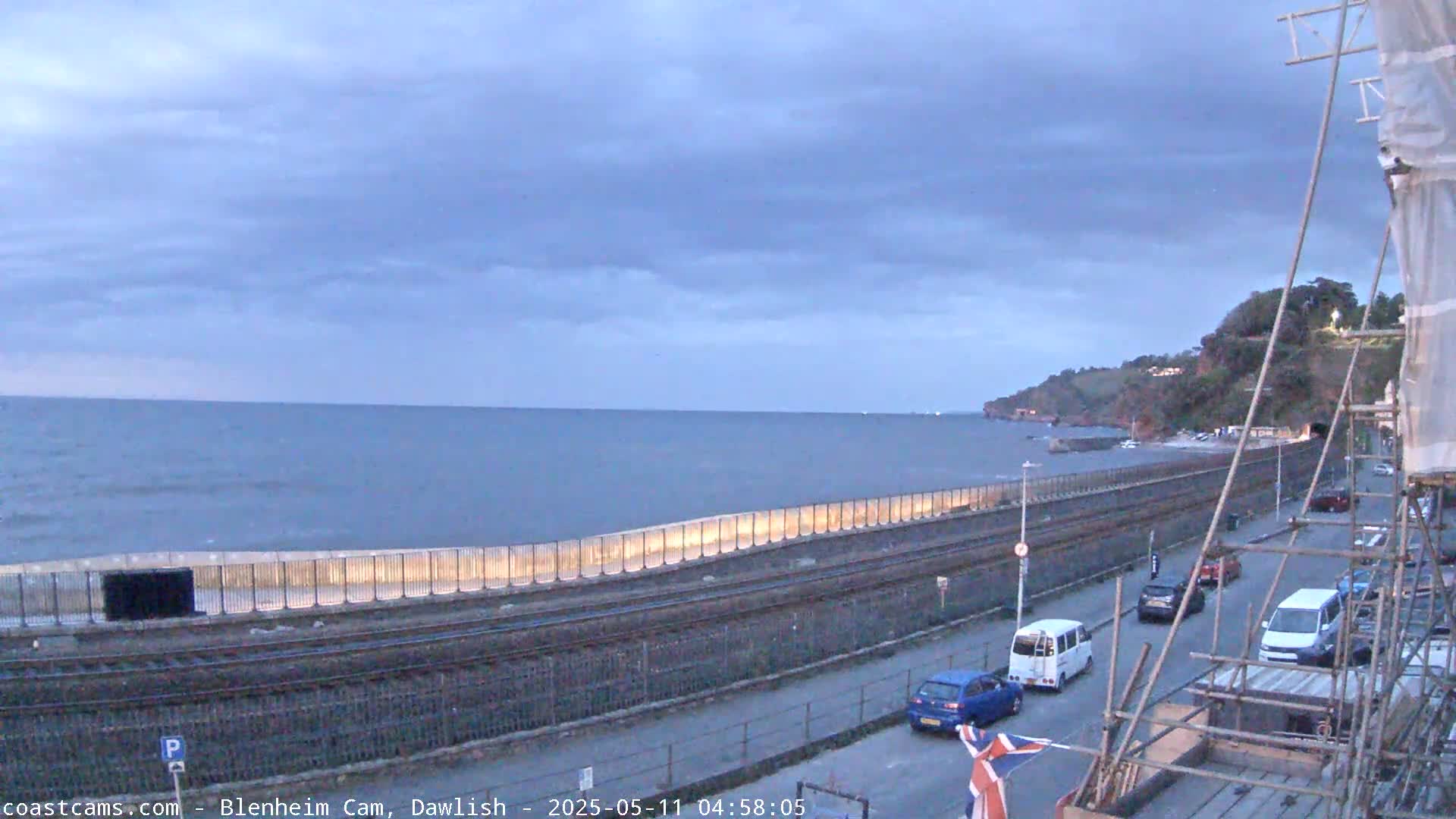 A calm sea stretches to a shoreline with a railway line, a road with cars, and a cliffside under an overcast sky.