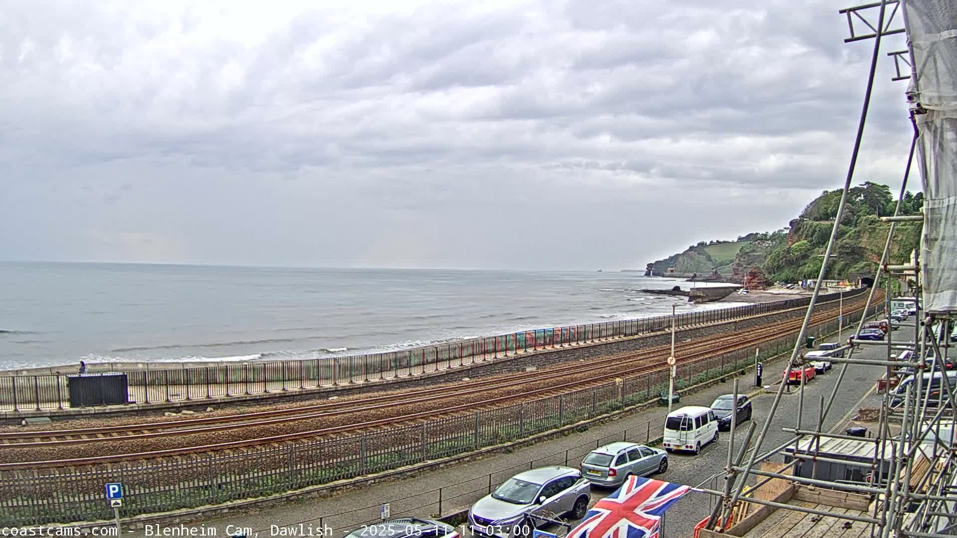 A coastal scene on an overcast day shows a beach, railroad tracks, parked cars, and a partially obscured building under construction.