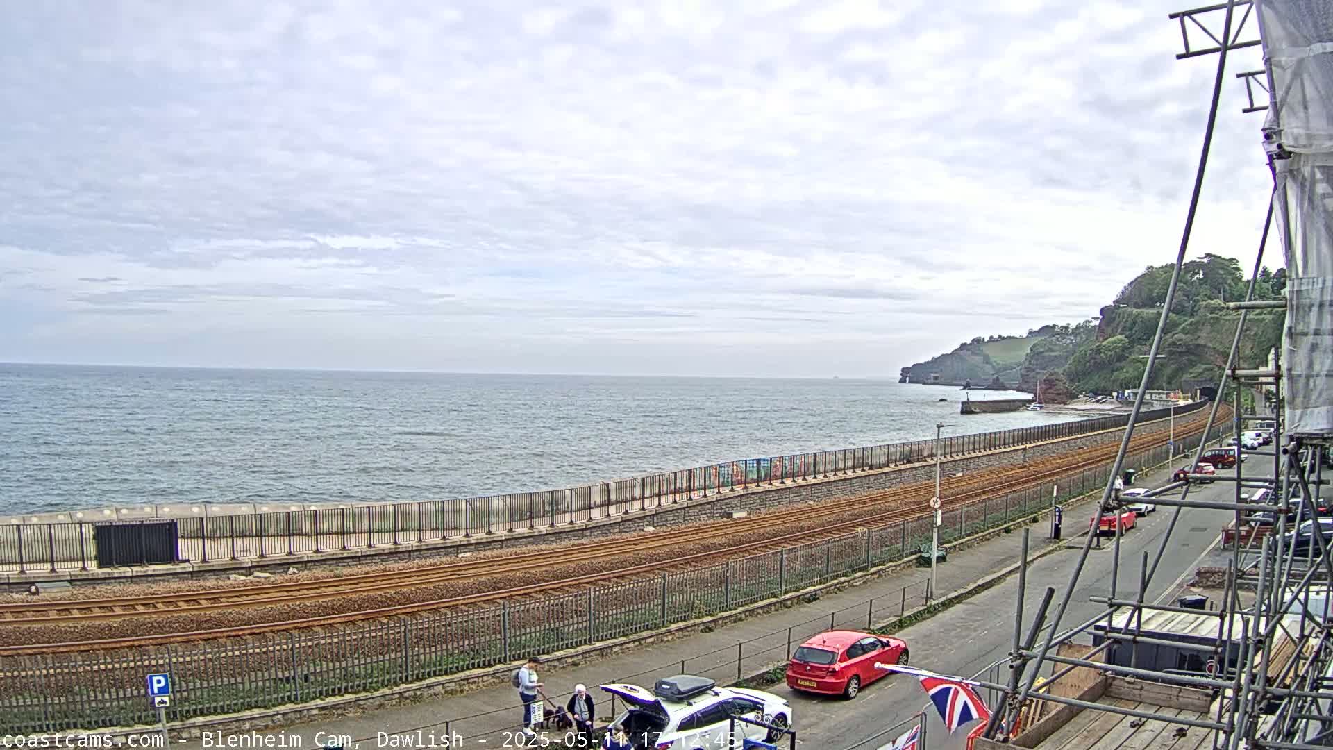 A partly cloudy day shows a coastal railway line running alongside a sea wall, with cars parked on a street next to construction scaffolding and a few people walking by.