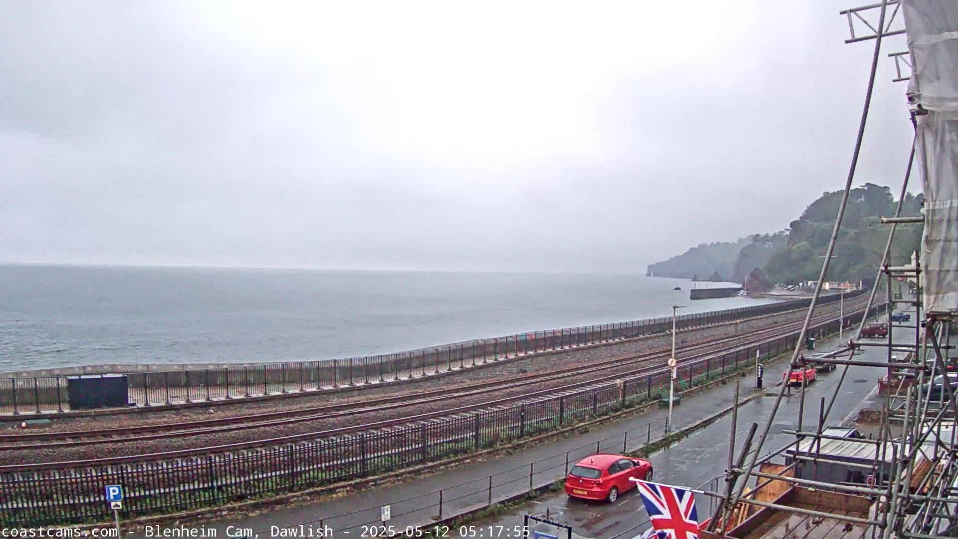 A seaside view on an overcast day shows a section of train tracks running alongside a coastal road, with a red car parked nearby and construction scaffolding visible in the foreground.