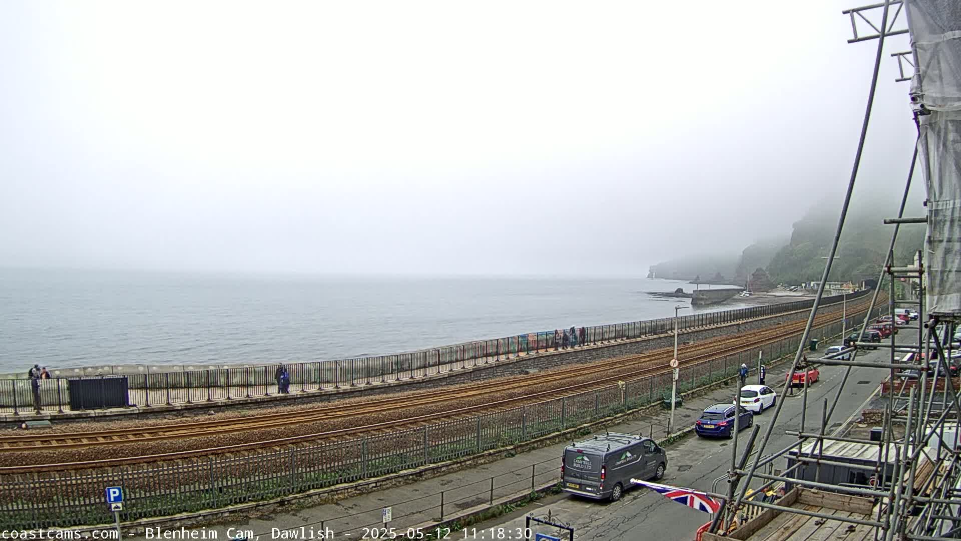 A foggy coastal scene shows train tracks running alongside a sea wall with several people and cars visible, and construction scaffolding in the foreground.