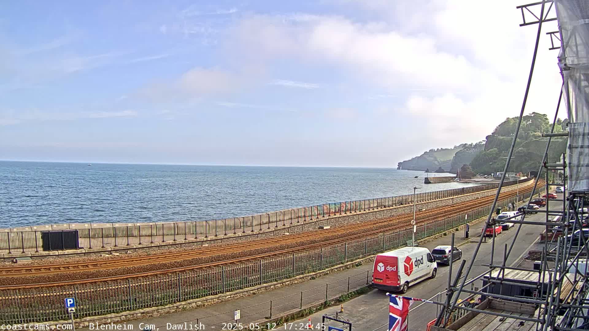 A mostly sunny day overlooking a calm ocean with a railway line running alongside a seaside road, where several cars and a delivery van are parked.
