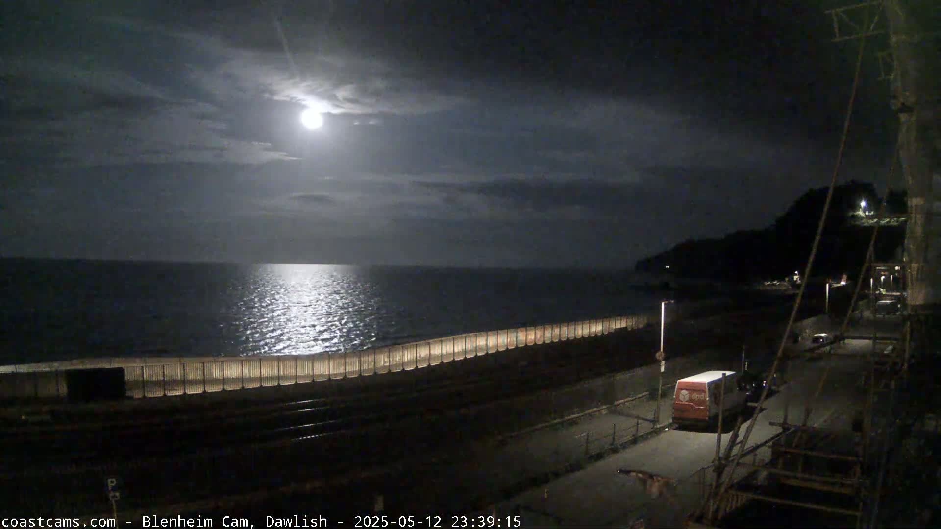 A bright moon illuminates a calm sea at night, seen from a coastal vantage point overlooking a seawall, a road, and a van.