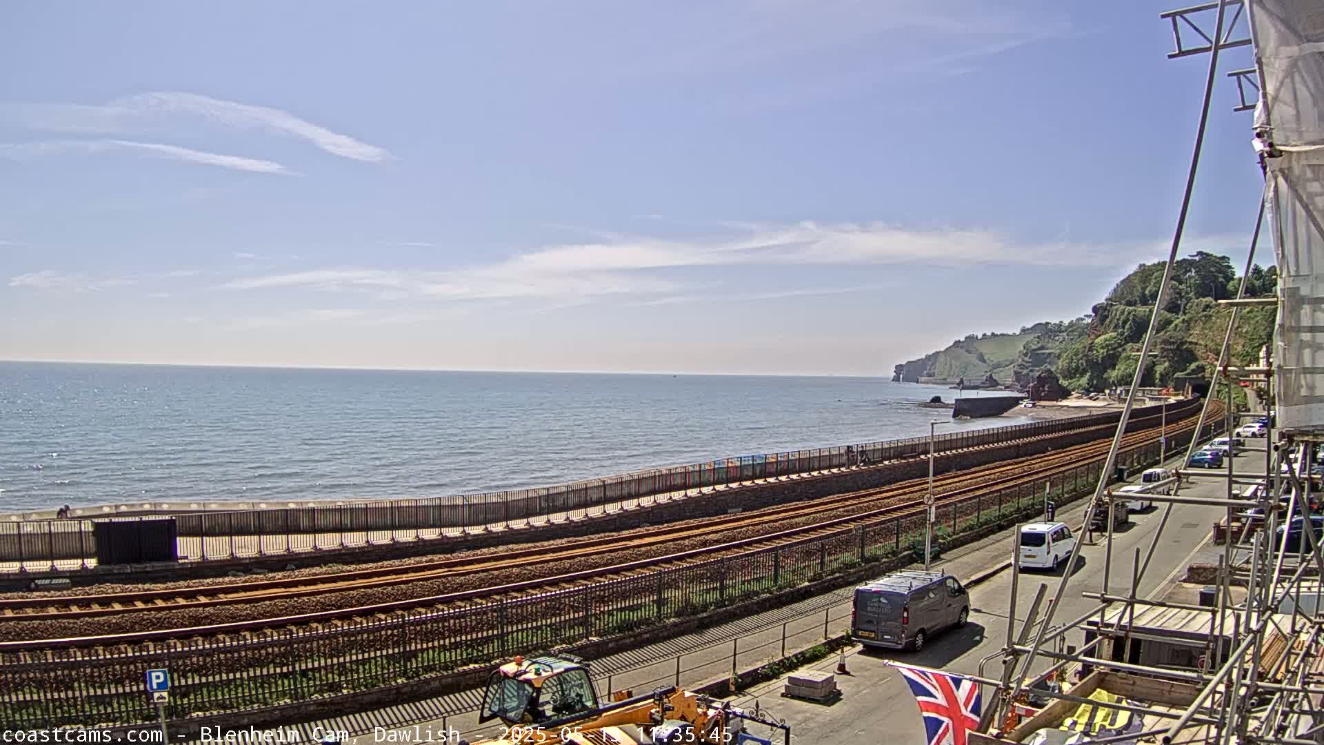 A coastal railway line runs alongside a calm sea under a mostly sunny sky, with construction scaffolding and vehicles visible along a nearby road.